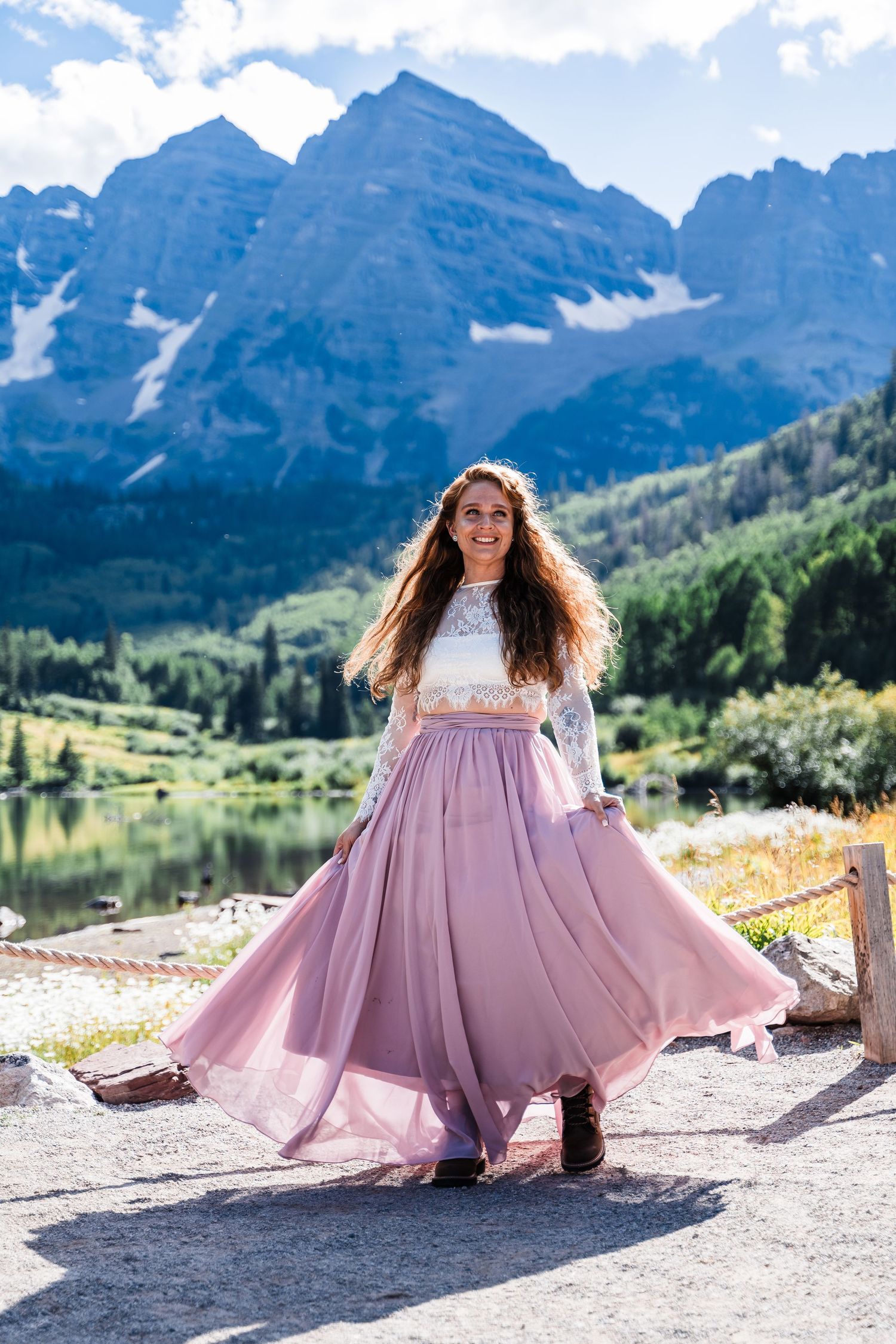 A person in a flowing pink dress twirls joyfully against a backdrop of majestic mountain peaks and a serene lake.