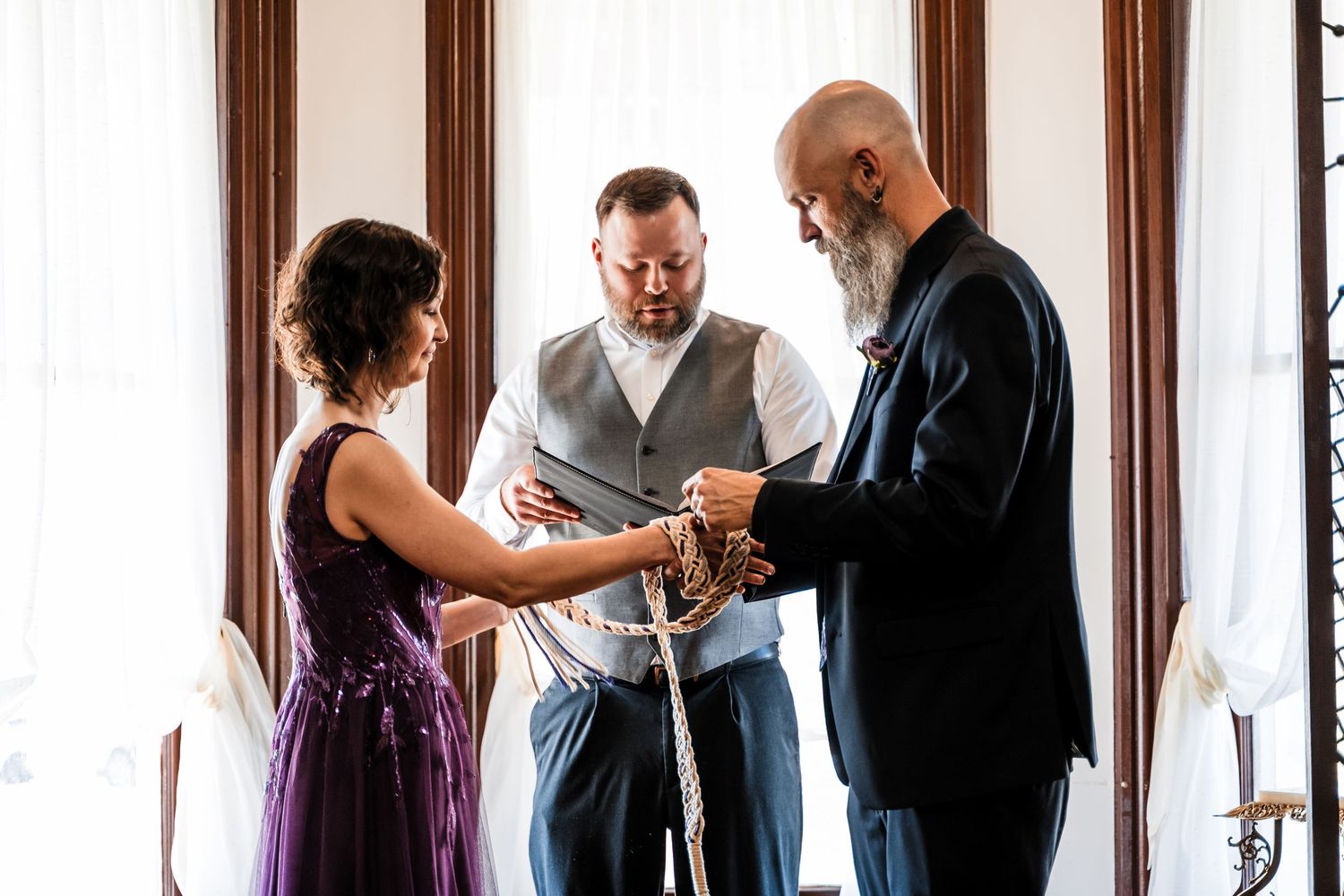 Couple performs handfasting ceremony with officiant in front of bright window.