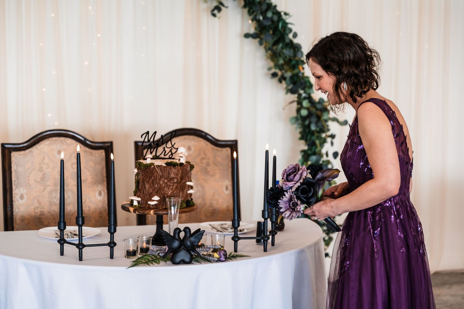 Bride in purple sequin gown arranges bouquet near rustic wedding cake with black candles.