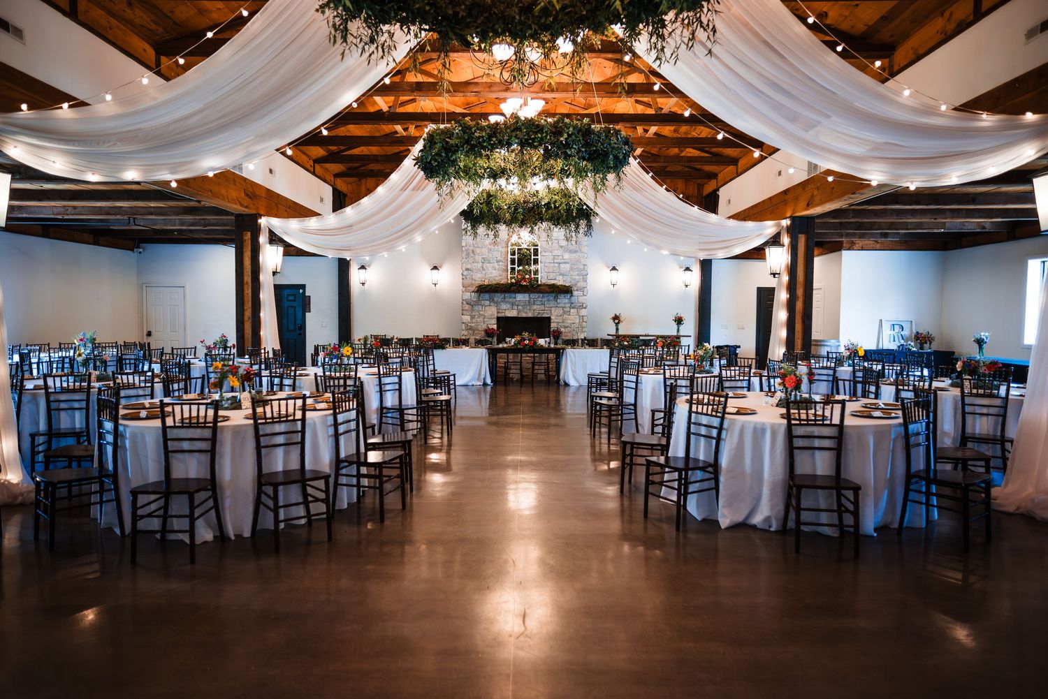 Elegant rustic wedding reception hall with white draped ceilings, round tables, and wooden beam accents.