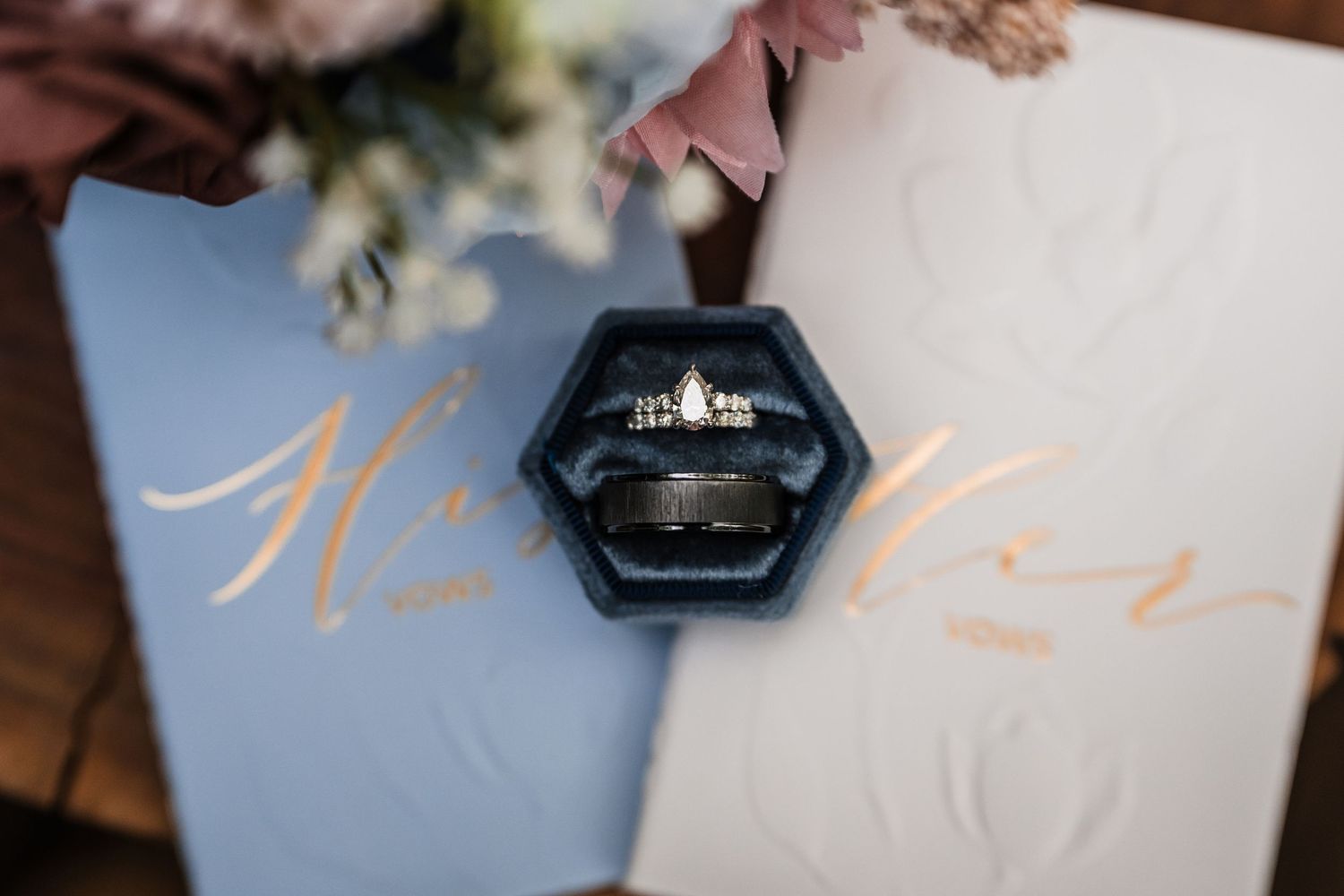Wedding rings displayed in a black hexagonal box on top of a light blue paper with rose gold script text.
