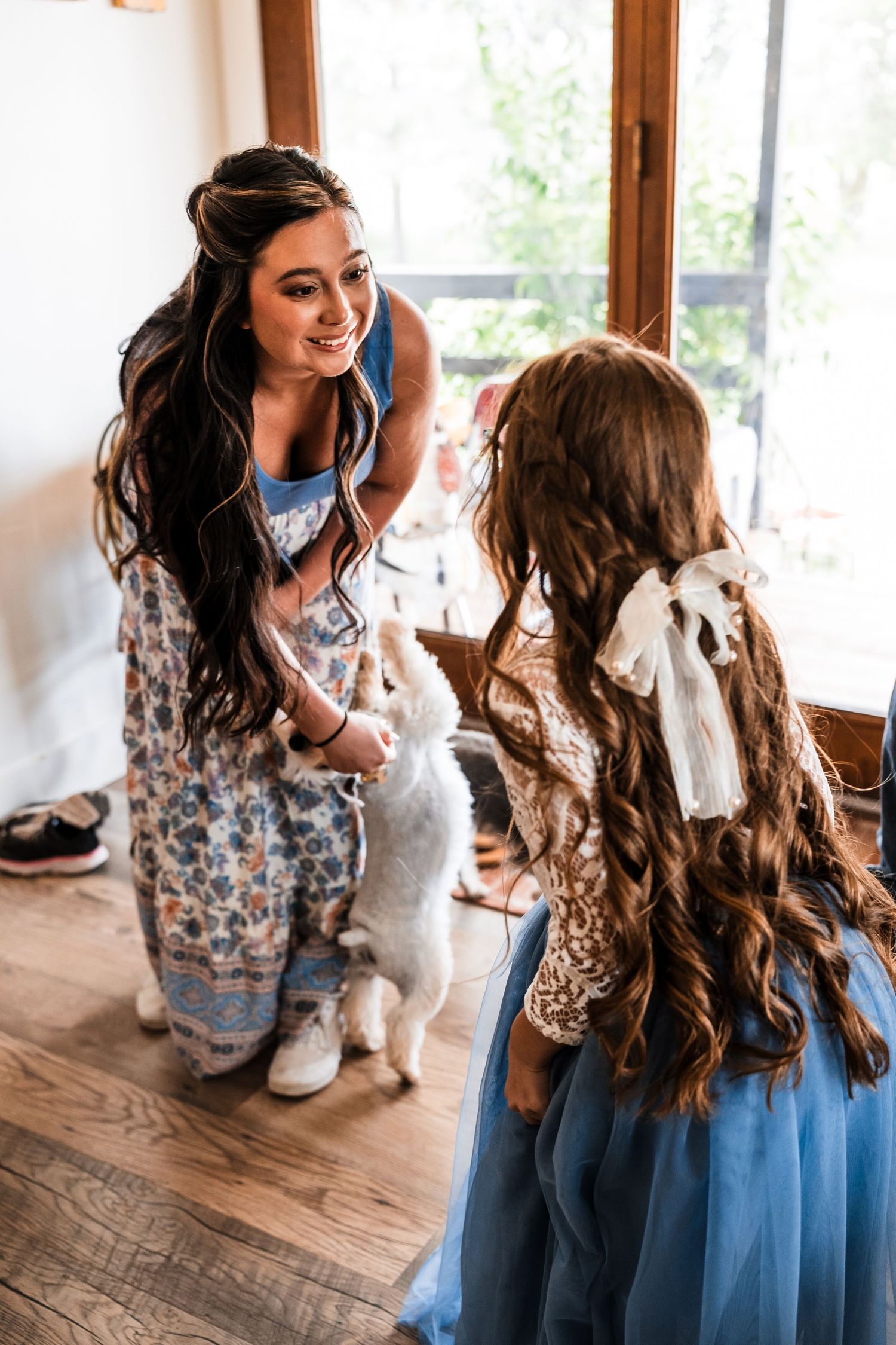Smiling bride in floral dress kneeling to interact with young girl in blue tulle skirt holding a white puppy.