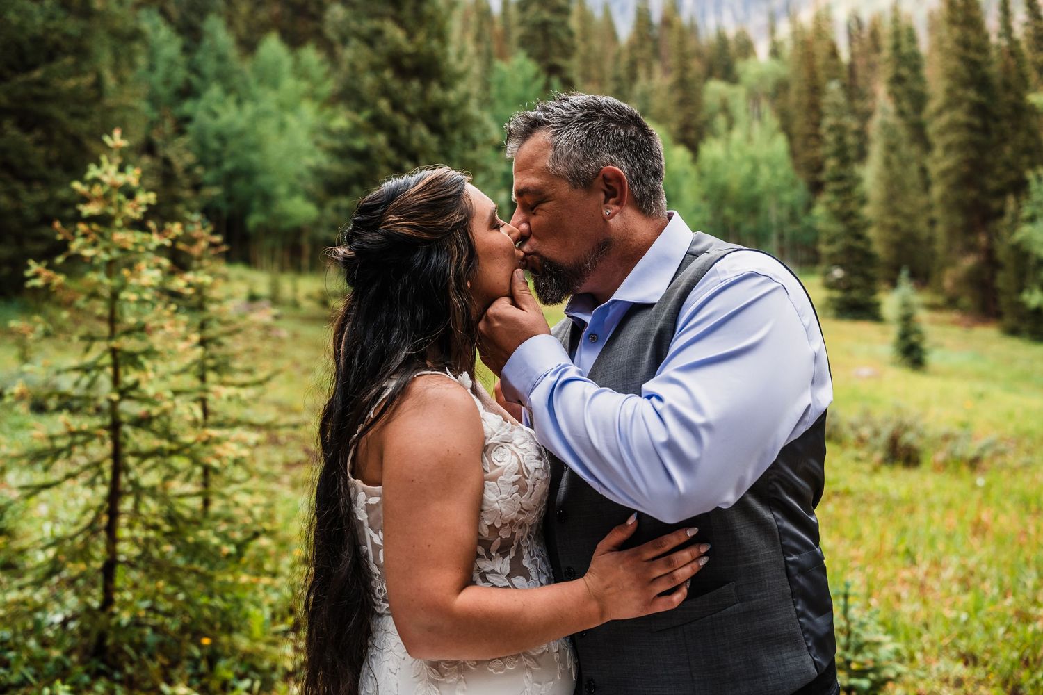 Couple sharing a romantic kiss in a lush green meadow surrounded by pine trees.