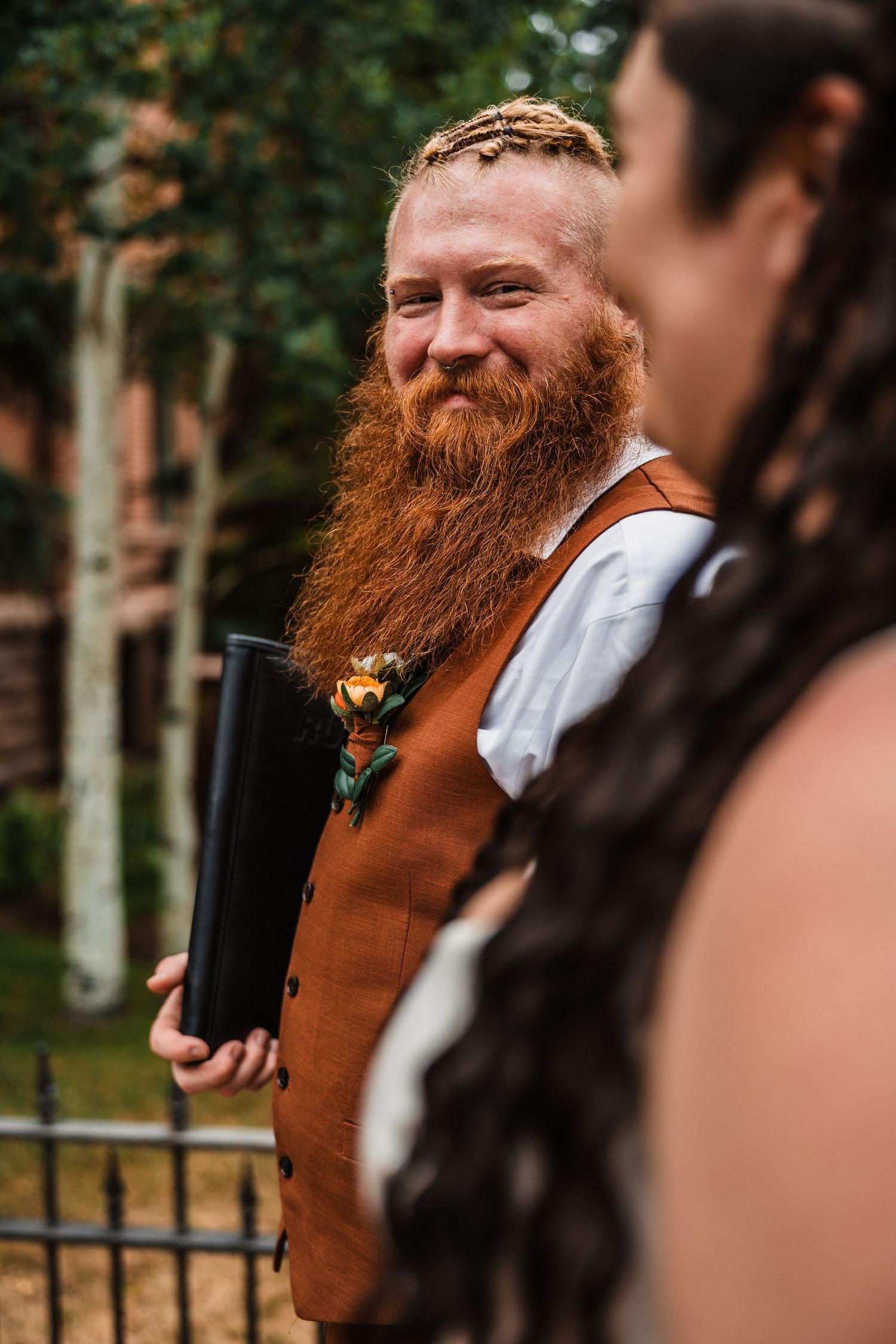 Smiling red-bearded groom in brown vest holds ceremony book during outdoor elopement.