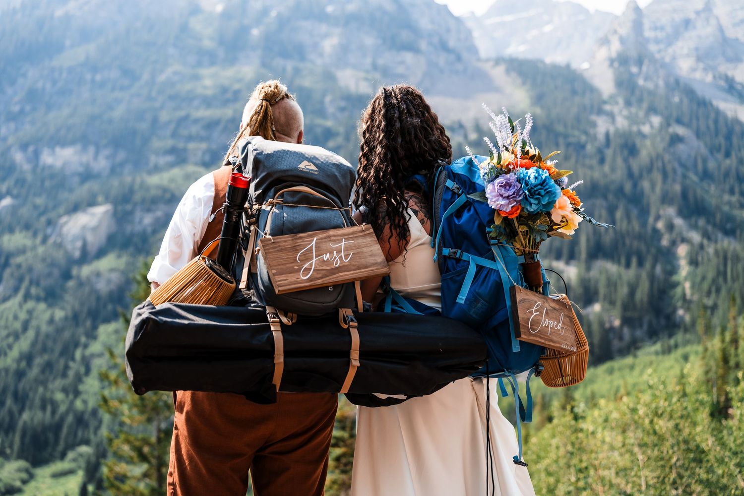 Two newlyweds with Just Married backpacks hold a colorful bouquet while overlooking a stunning mountain forest landscape.