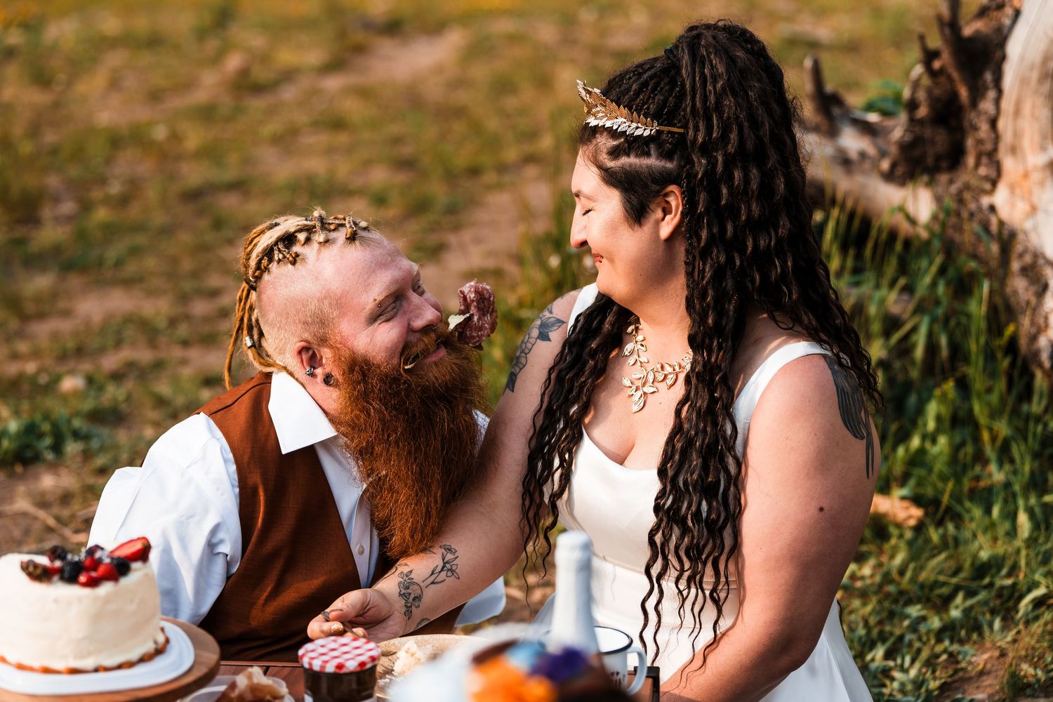 Bearded groom with red braids and tattooed bride with dreadlocks share a joyful moment at outdoor wedding table.