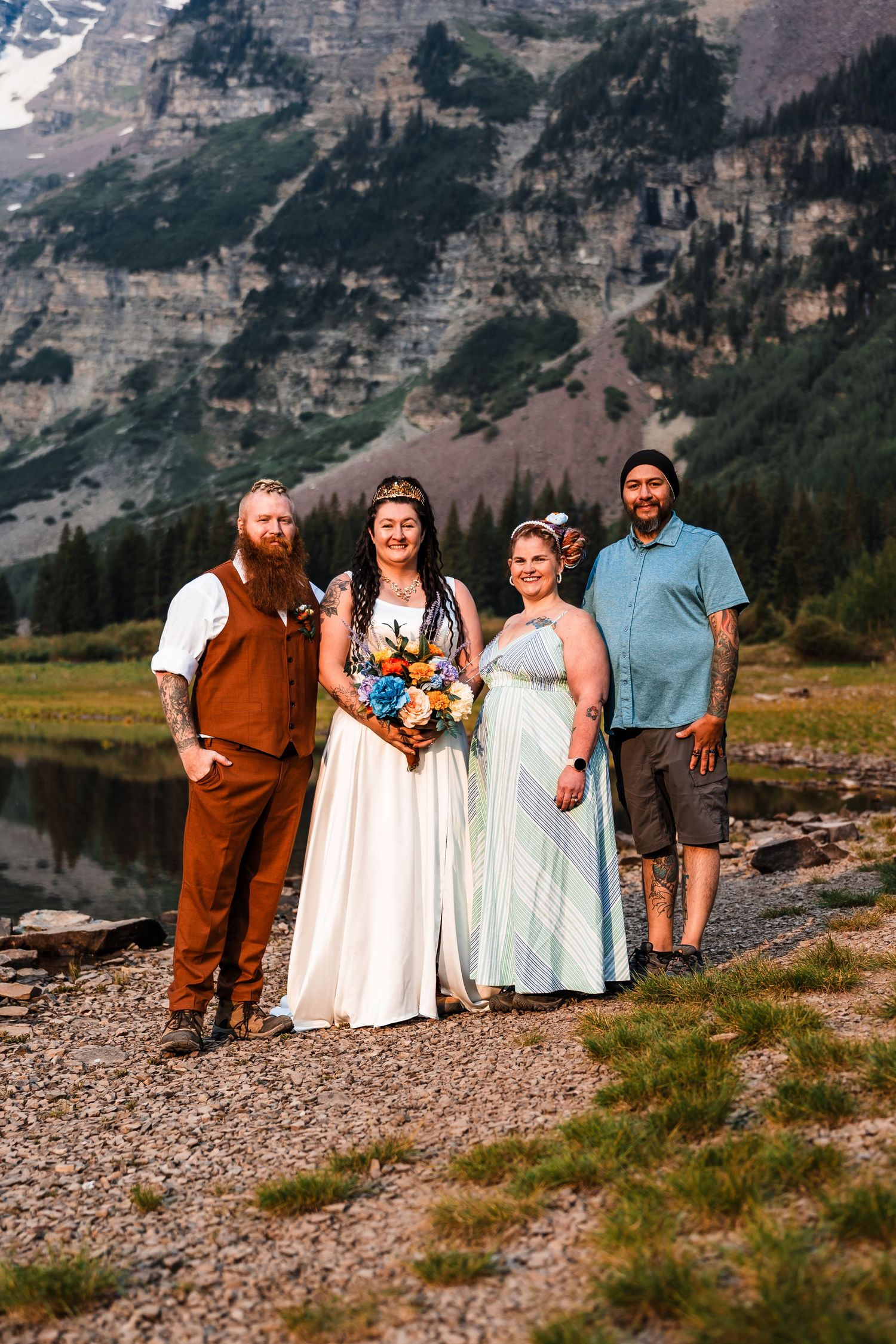 Wedding party of four posing near a mountain lake with rocky peaks and pine trees in the background.