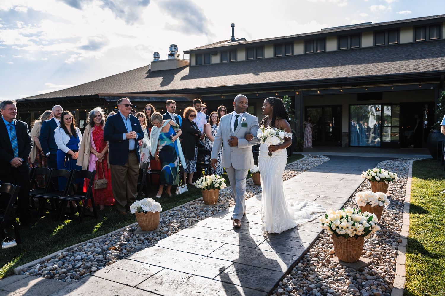 Bride and groom walk down outdoor aisle surrounded by guests at elegant wedding venue under blue sky.