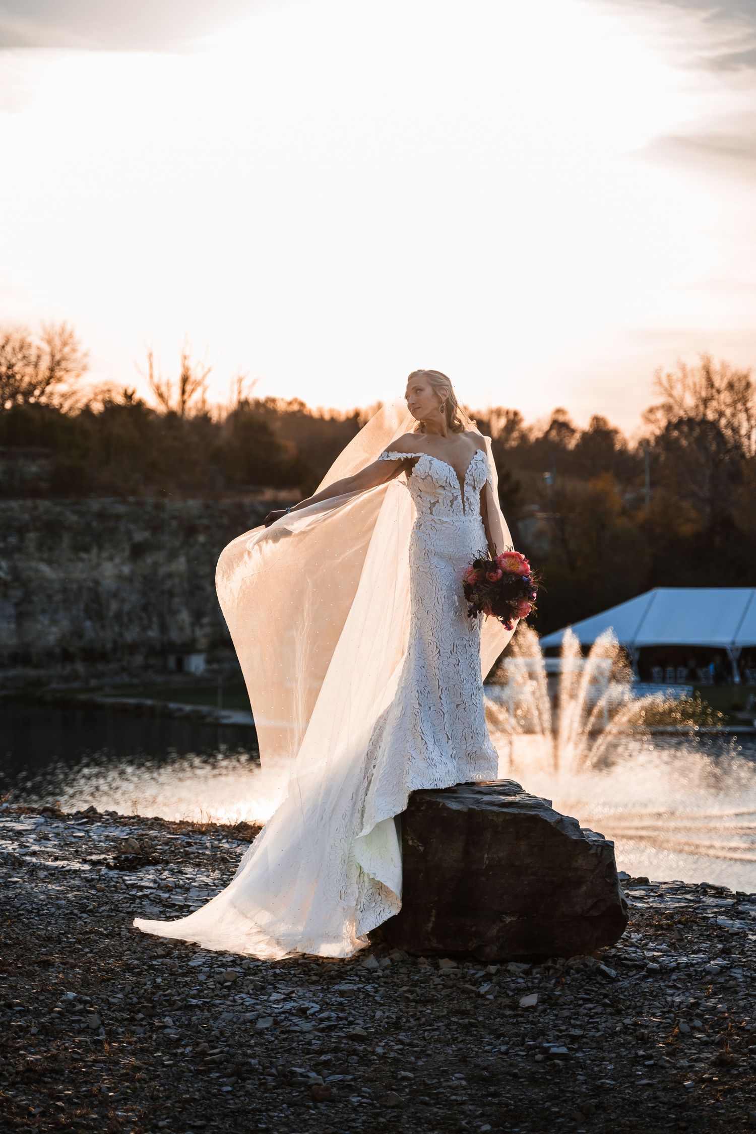 A bride in a flowing white dress stands gracefully on a rocky shoreline at sunset, her veil billowing in the wind.