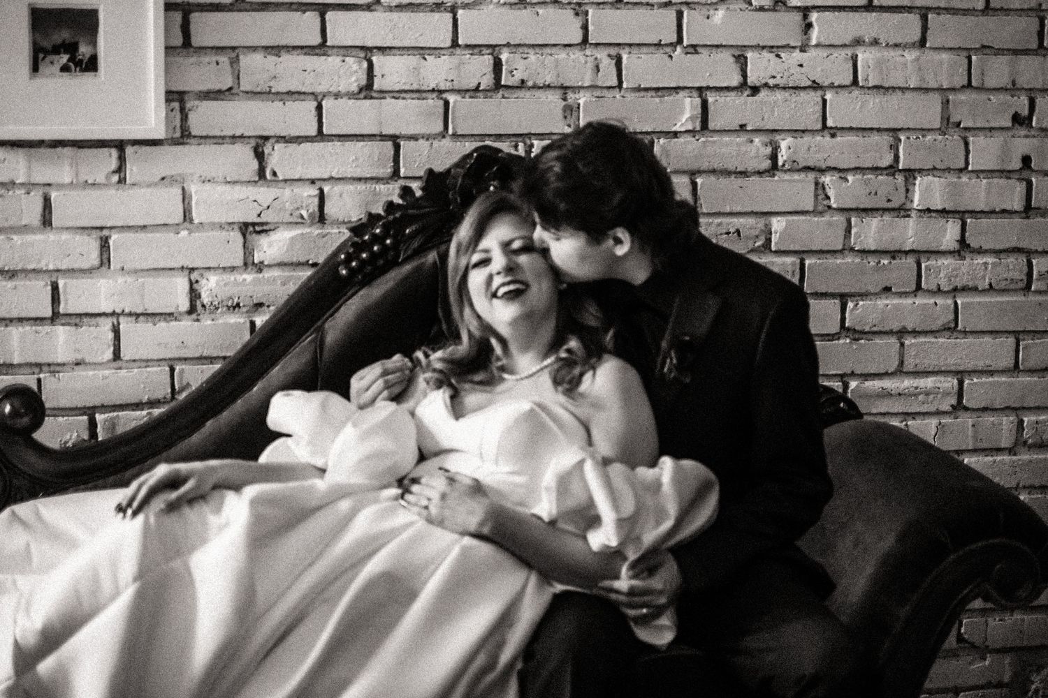 Intimate black and white moment of a couple sharing laughter on a couch against brick wall.