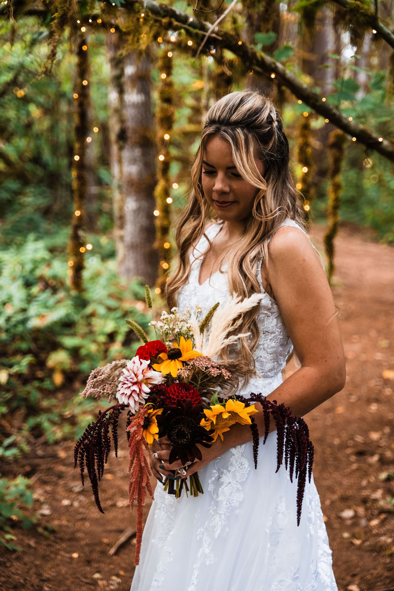 Bride holding vibrant fall-colored bouquet against forest backdrop with twinkling lights.