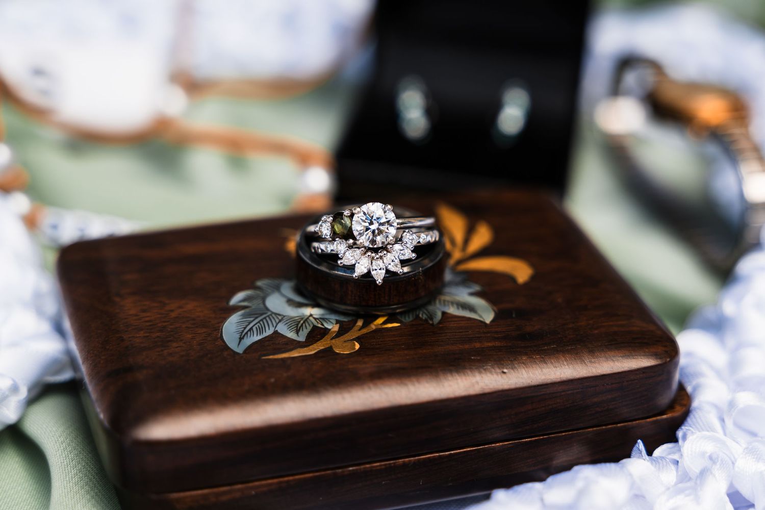 Diamond engagement ring and wedding bands rest on wooden box with floral detail.