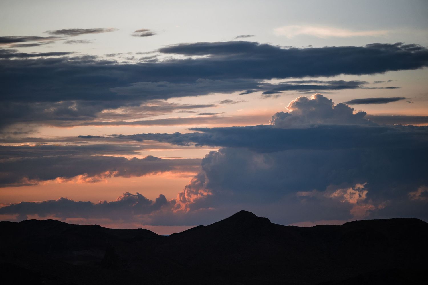Dramatic sunset sky with dark clouds silhouetting mountain peaks in orange and blue hues.