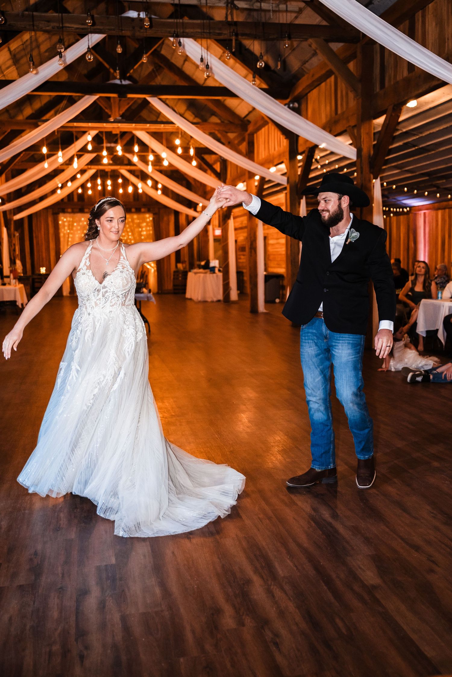A couple dancing in a rustic barn venue decorated with string lights and white fabric draping.