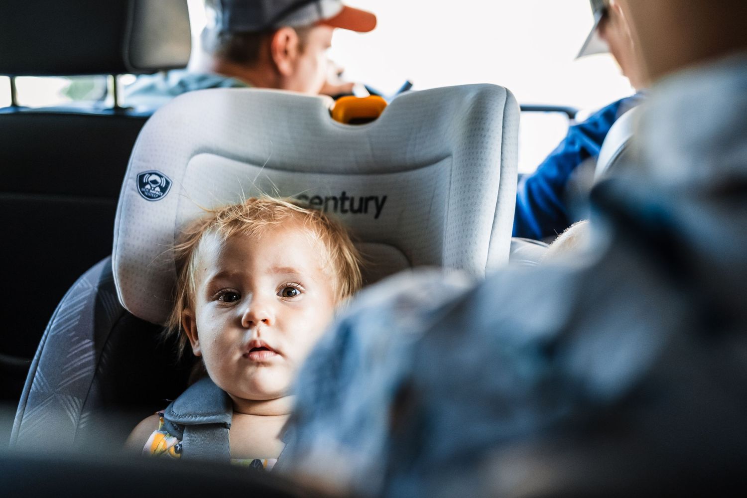 Young child sits in a Century brand car seat during a car ride, looking off to the side while being supervised.
