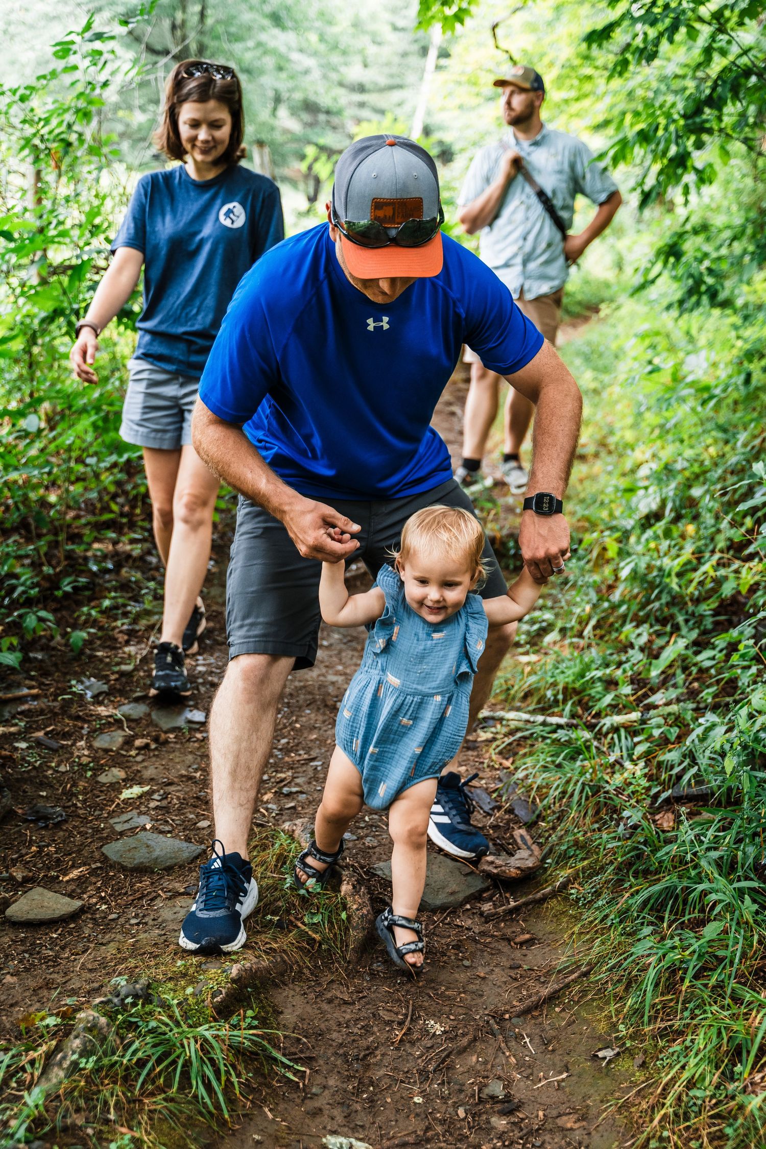 A group hiking on a forest trail in summer, with two adults and a small child learning to walk on the dirt path.