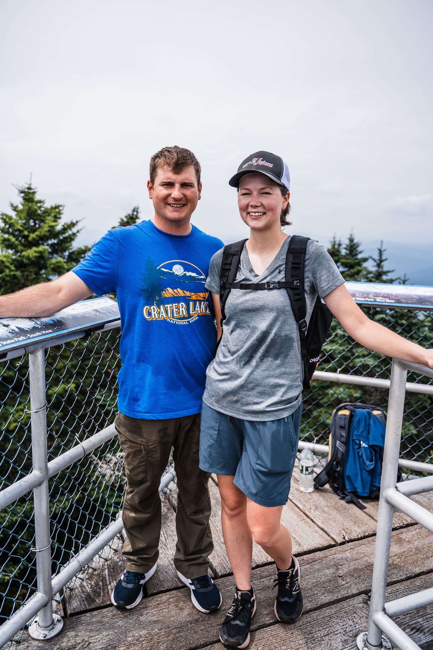 Two hikers pose together on a wooden observation deck surrounded by mountain views and evergreen trees.