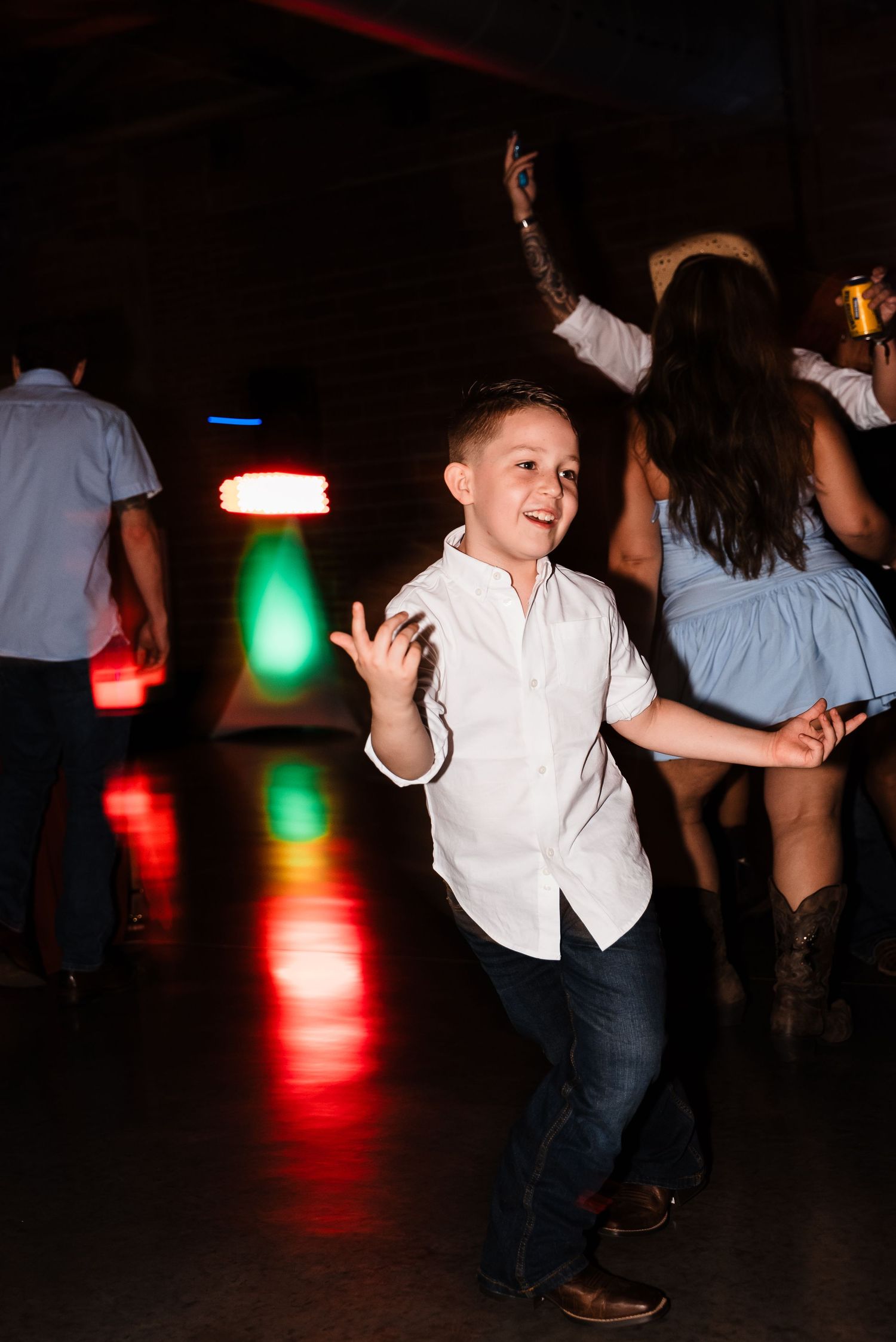 Young boy dancing joyfully on a dark dance floor with colorful lights in the background at an event.