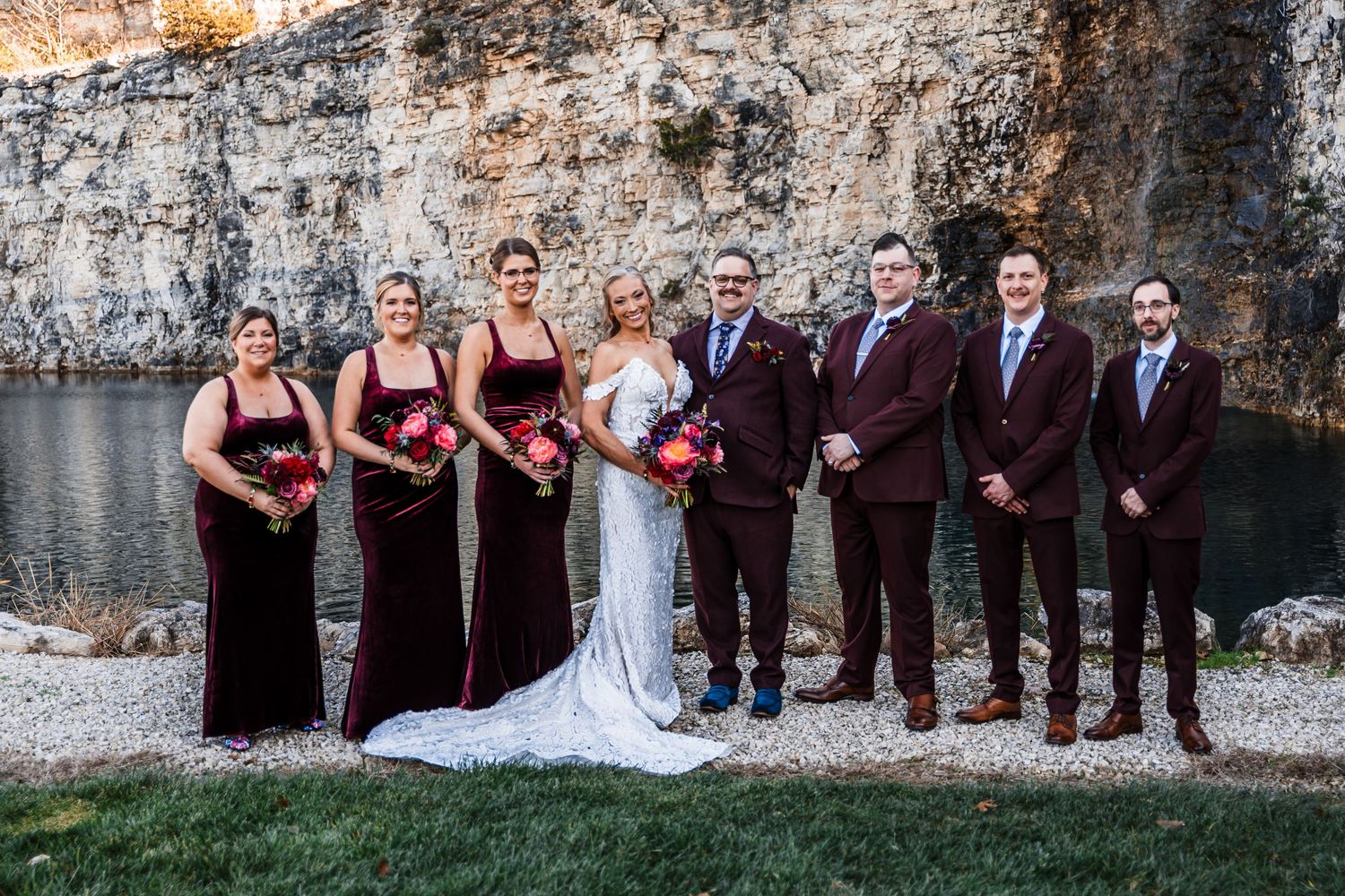 Wedding party in burgundy and formal attire poses against rustic stone wall for outdoor photos.