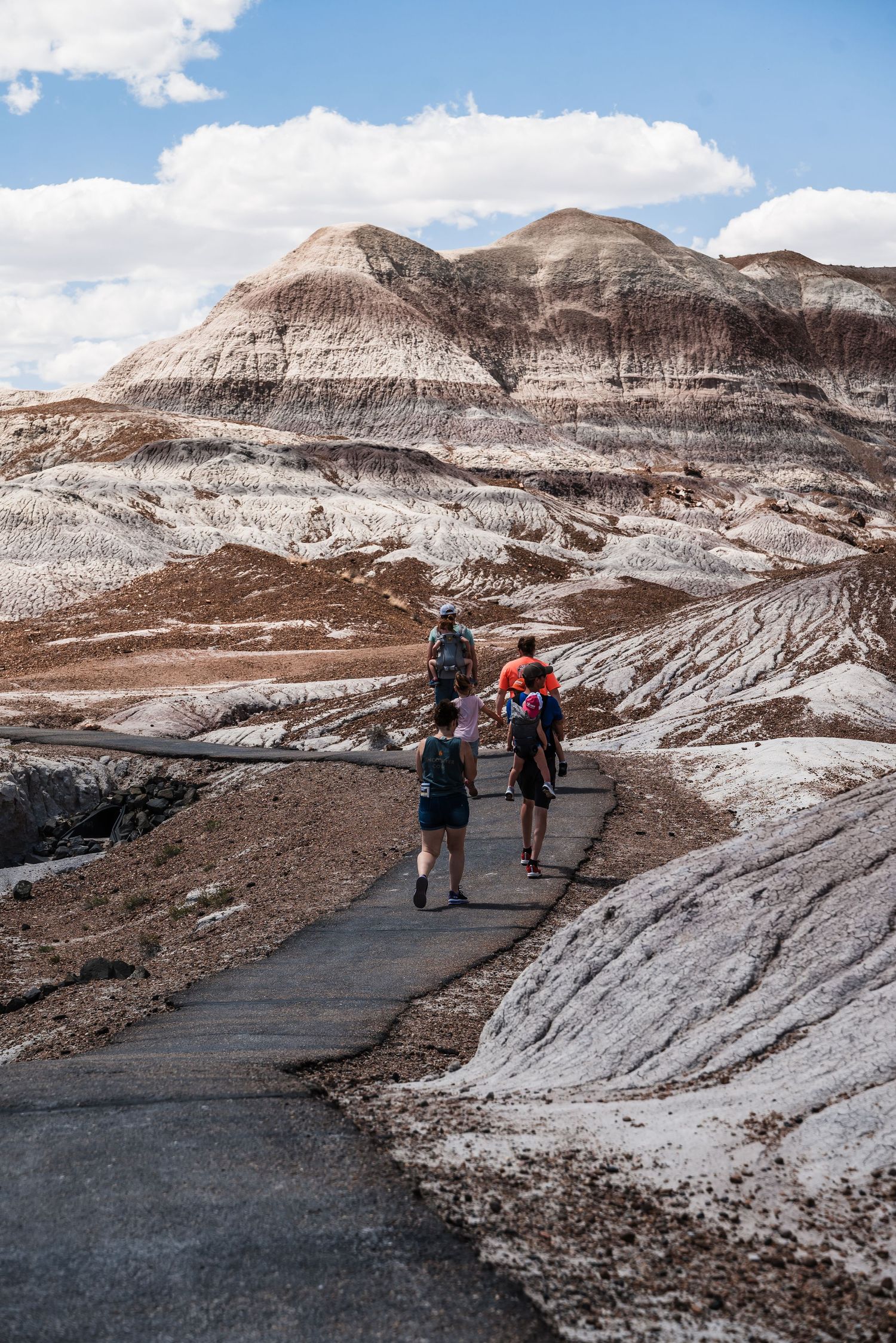 Hikers follow a paved trail through the striped badlands formations of a desert landscape under blue skies.