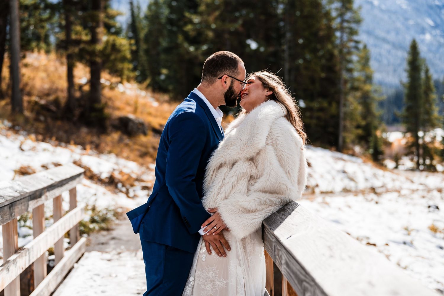 A couple shares romantic moments on a snowy mountain path surrounded by evergreen trees.