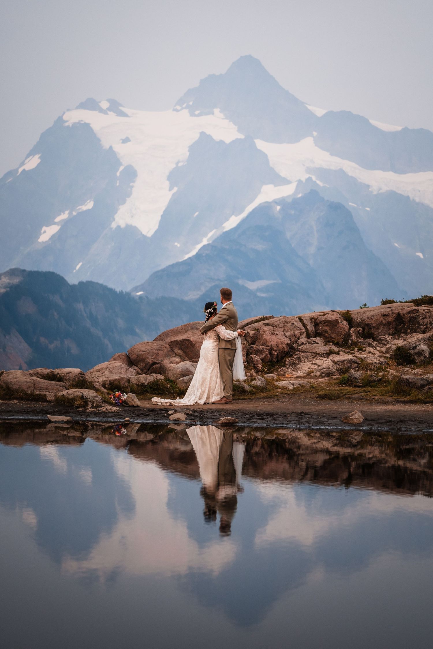 A couple stands reflected in calm mountain waters with dramatic snow-capped peaks in the background.
