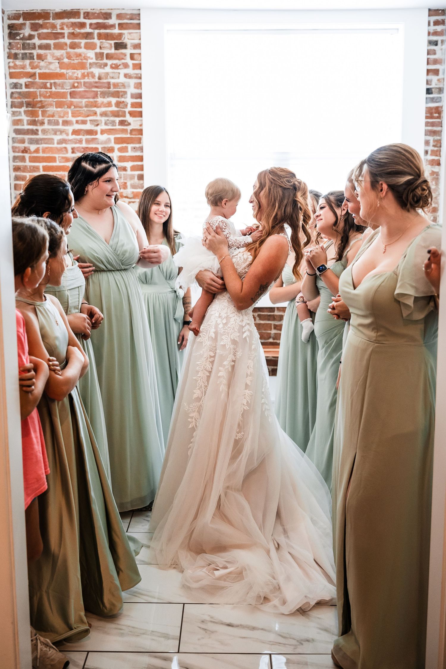 Bridesmaids in sage green dresses gather around a bride in a white gown against a brick wall backdrop.