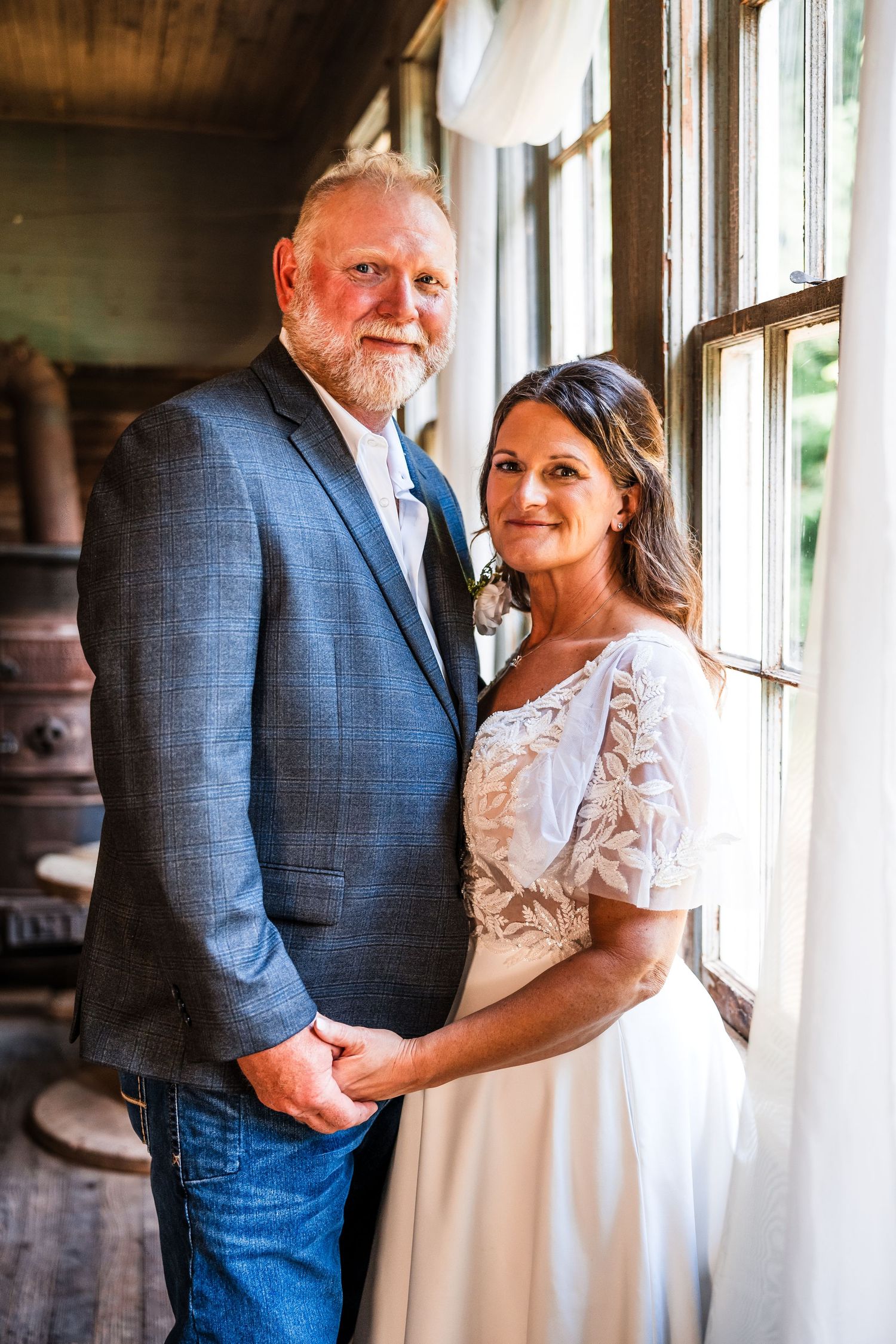 A couple shares an intimate embrace by large windows in a rustic indoor setting.