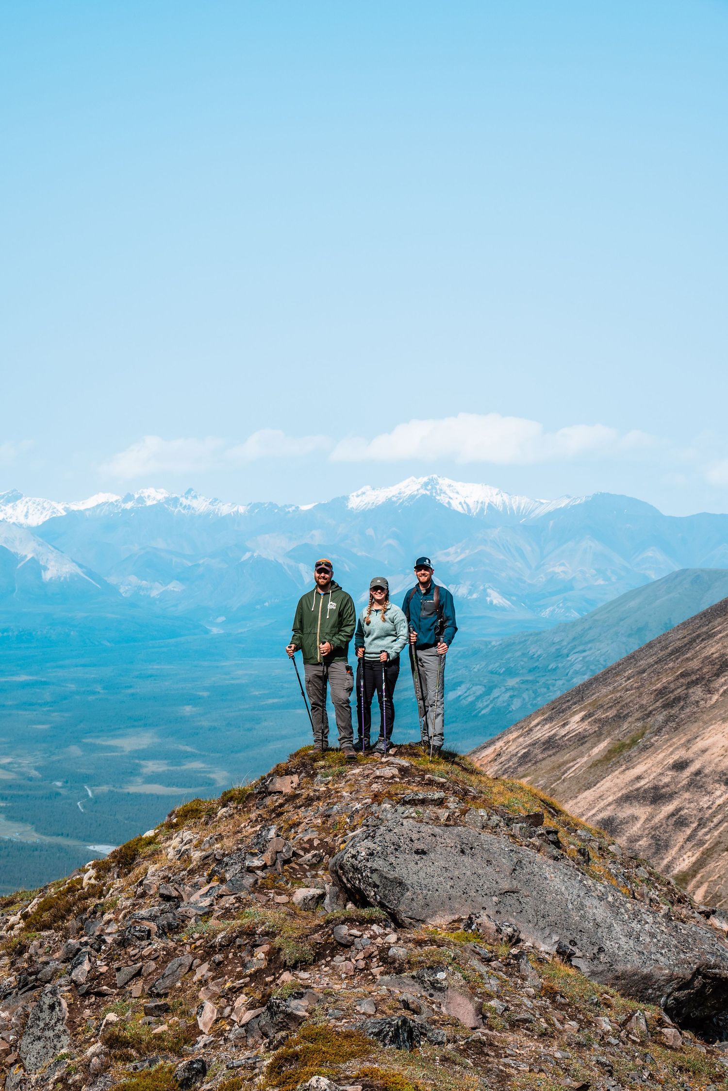 Three adventurers stand triumphantly on a rocky mountain peak overlooking snow-capped ranges and valleys below.