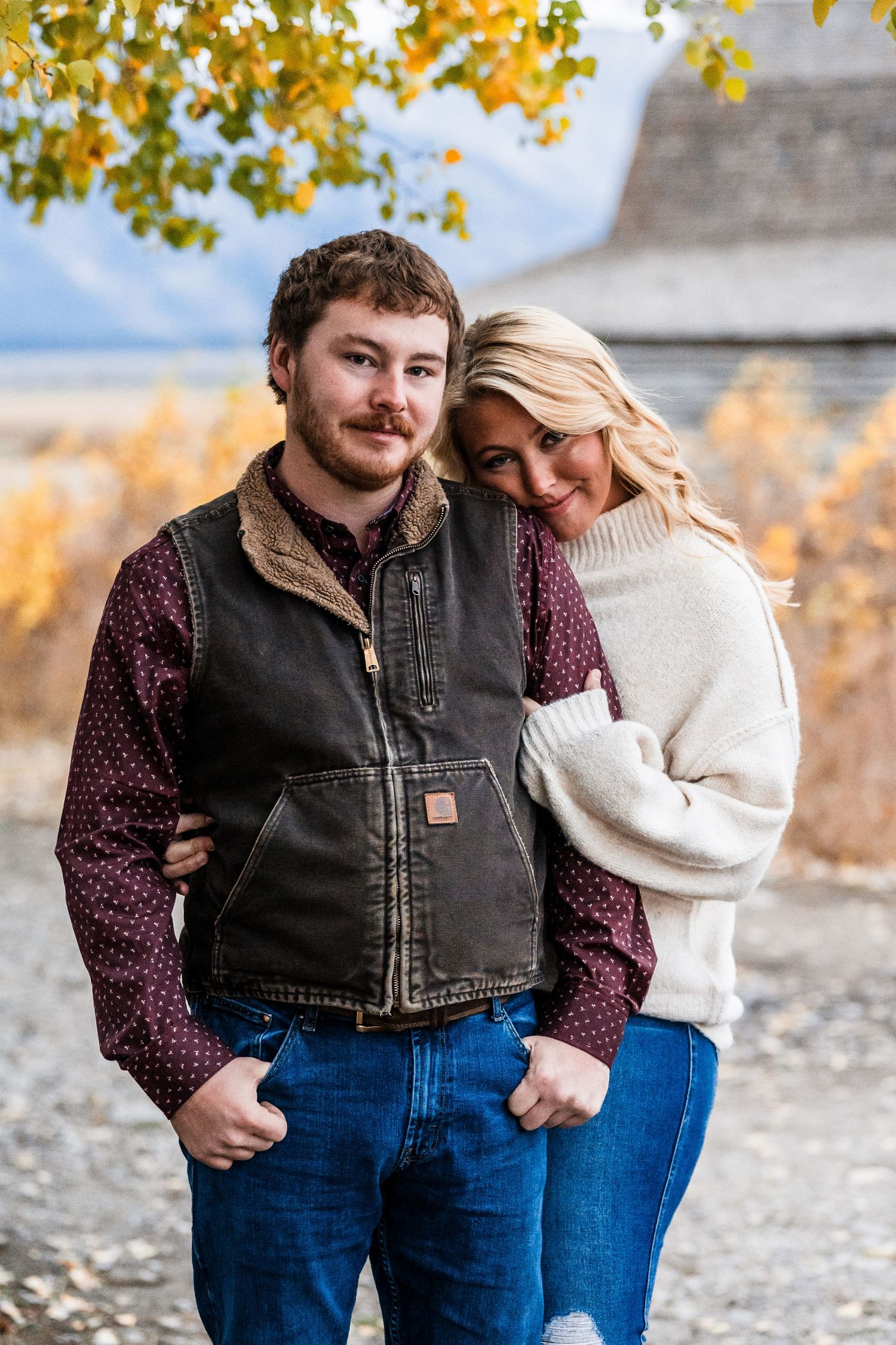 A couple in casual fall attire embraces against an autumn backdrop with golden leaves.