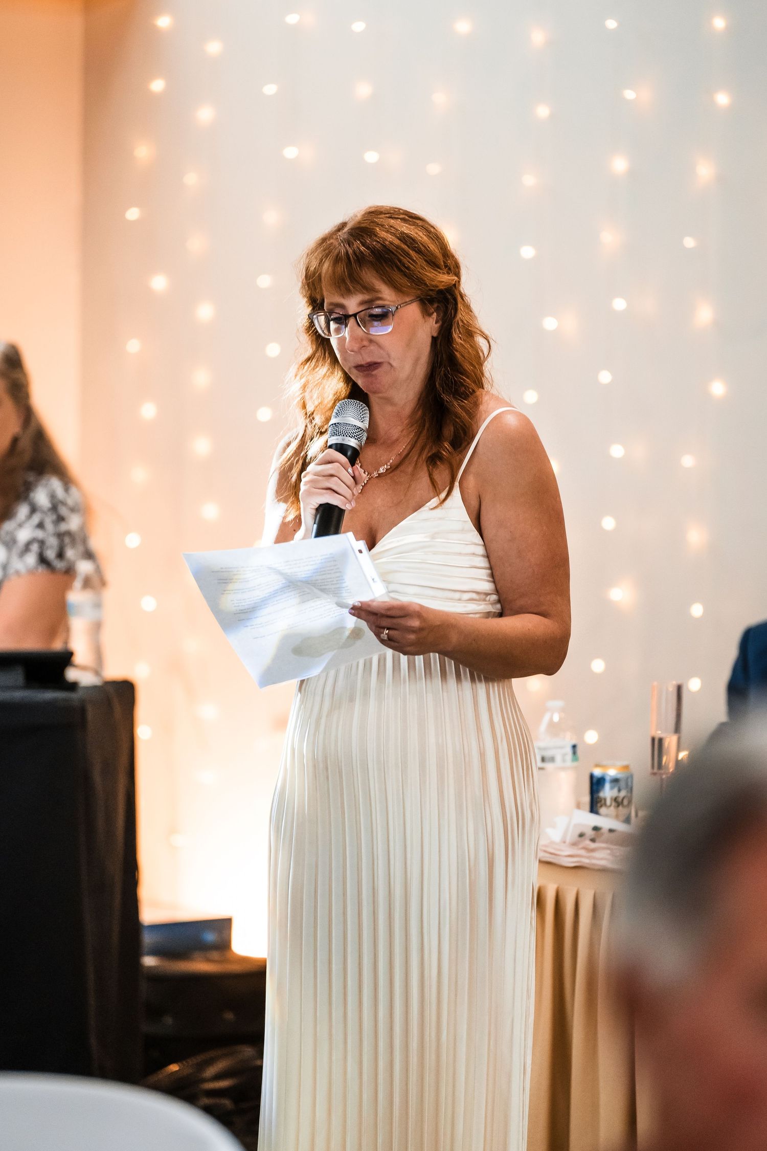 A person in a white pleated dress reads from papers at a wedding reception with twinkle lights in background.