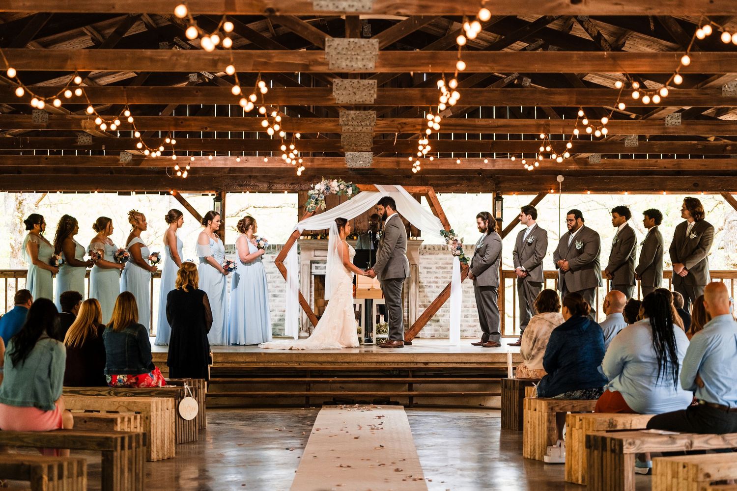 A rustic wooden barn venue decorated with string lights hosts an indoor wedding ceremony with guests seated on wooden pews.