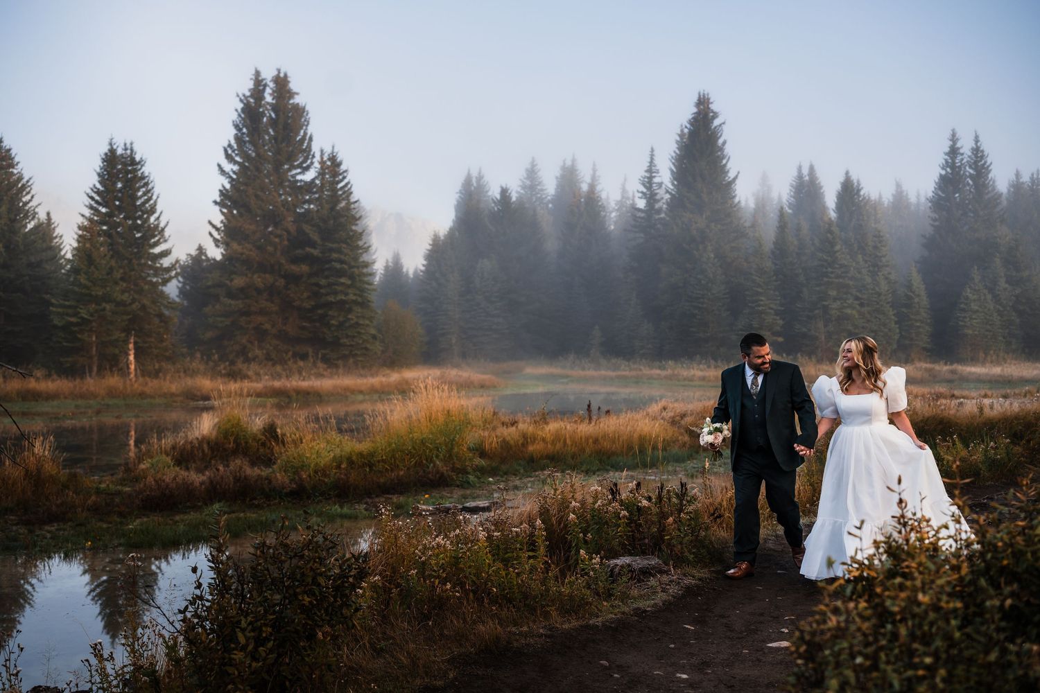 Wedding couple walking along a serene lake shore with pine trees and mountains in the background.