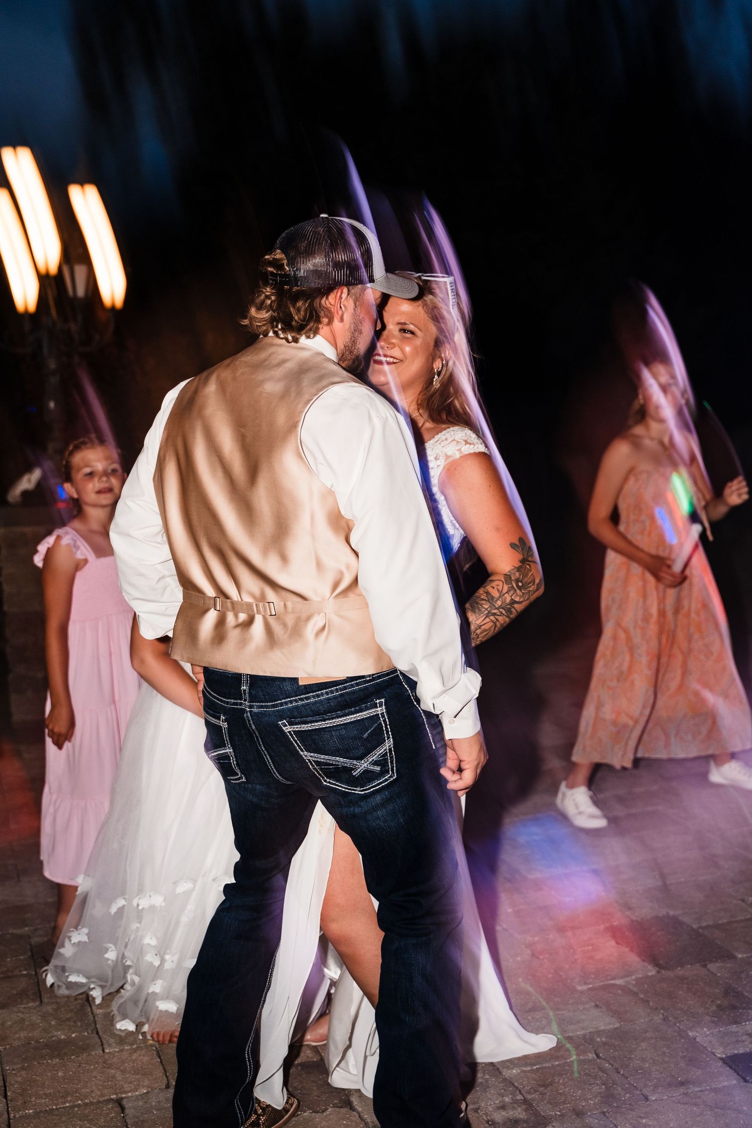 A couple dances at their wedding reception while guests in pink dresses celebrate in the background.