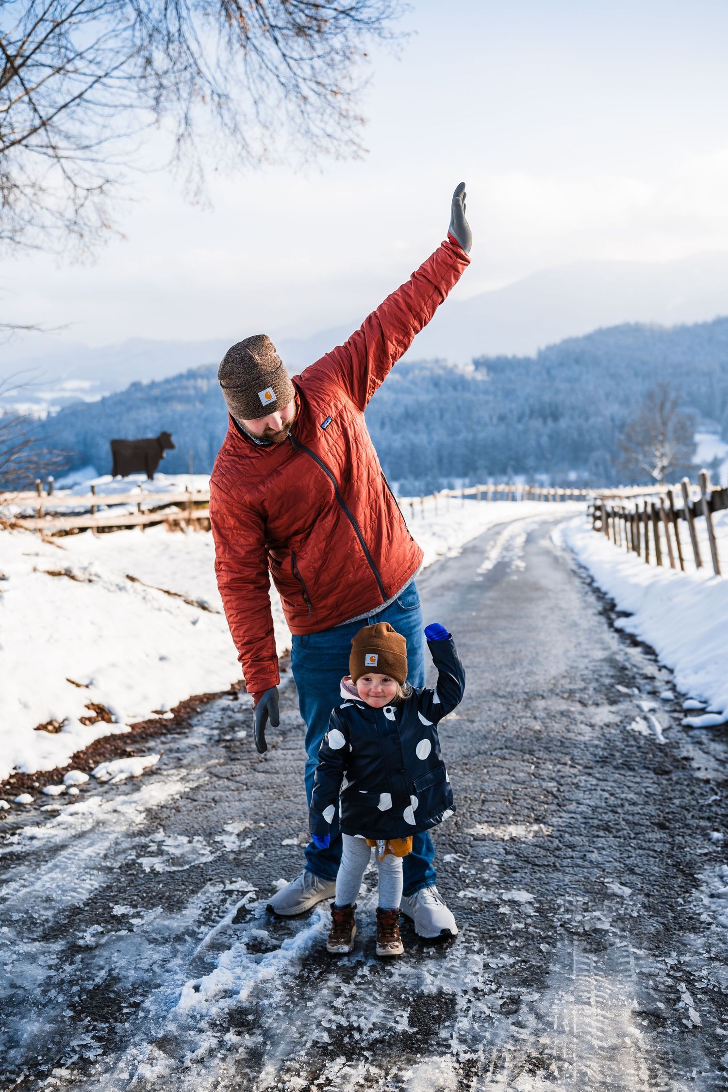Parent and child in red winter jacket playing together on a snowy mountain path surrounded by wooden fences.