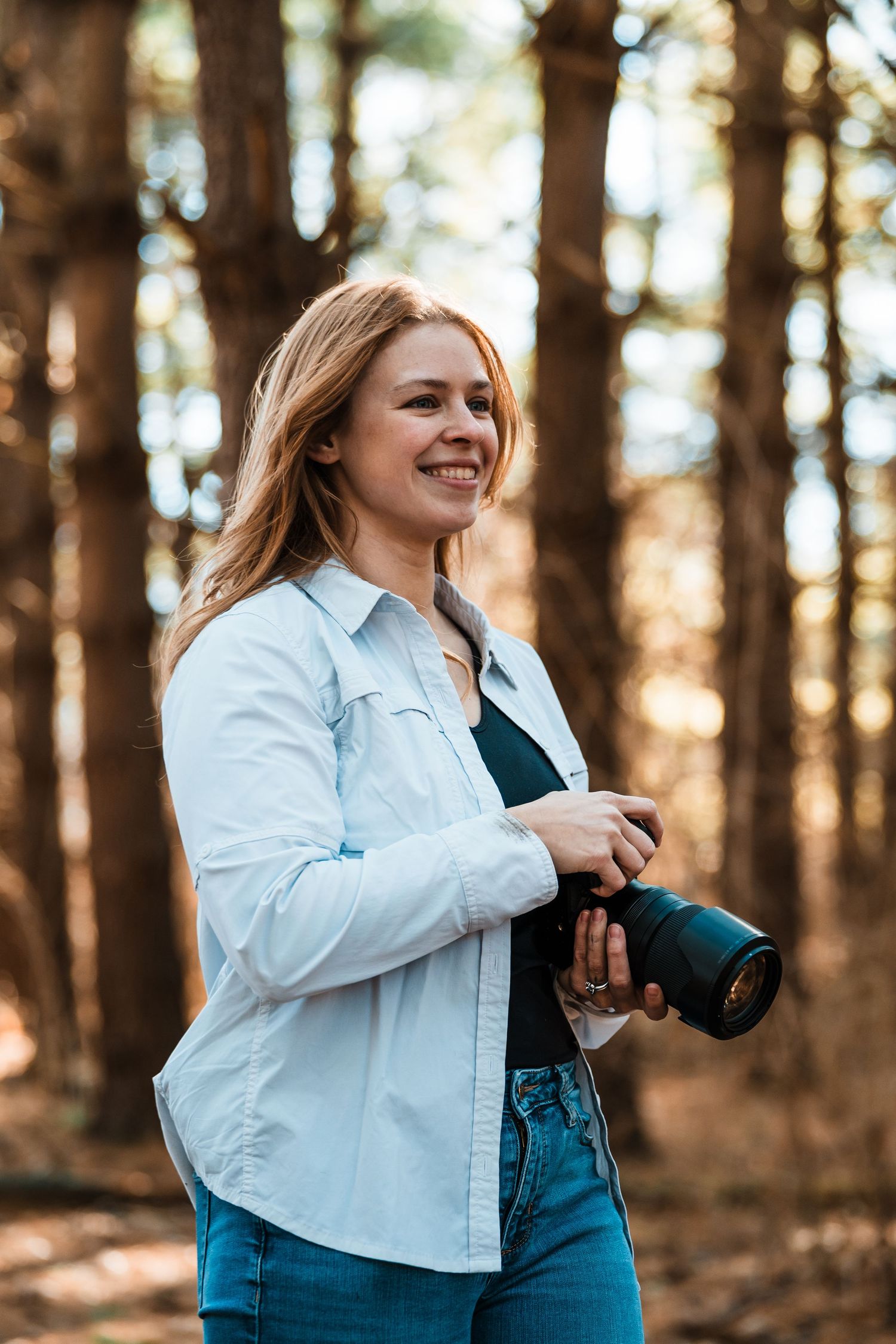 Photographer in white shirt and jeans works with camera equipment in a forest setting.