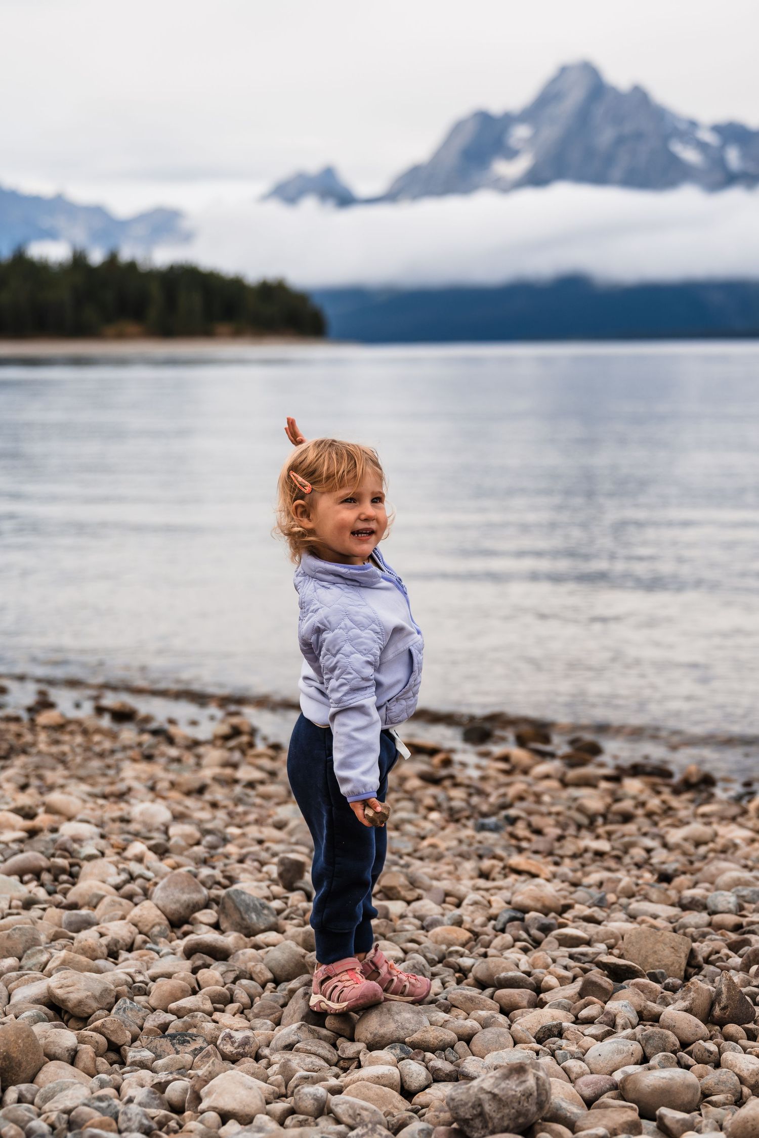 Small child explores rocky shoreline with snow-capped mountains and lake in the background.