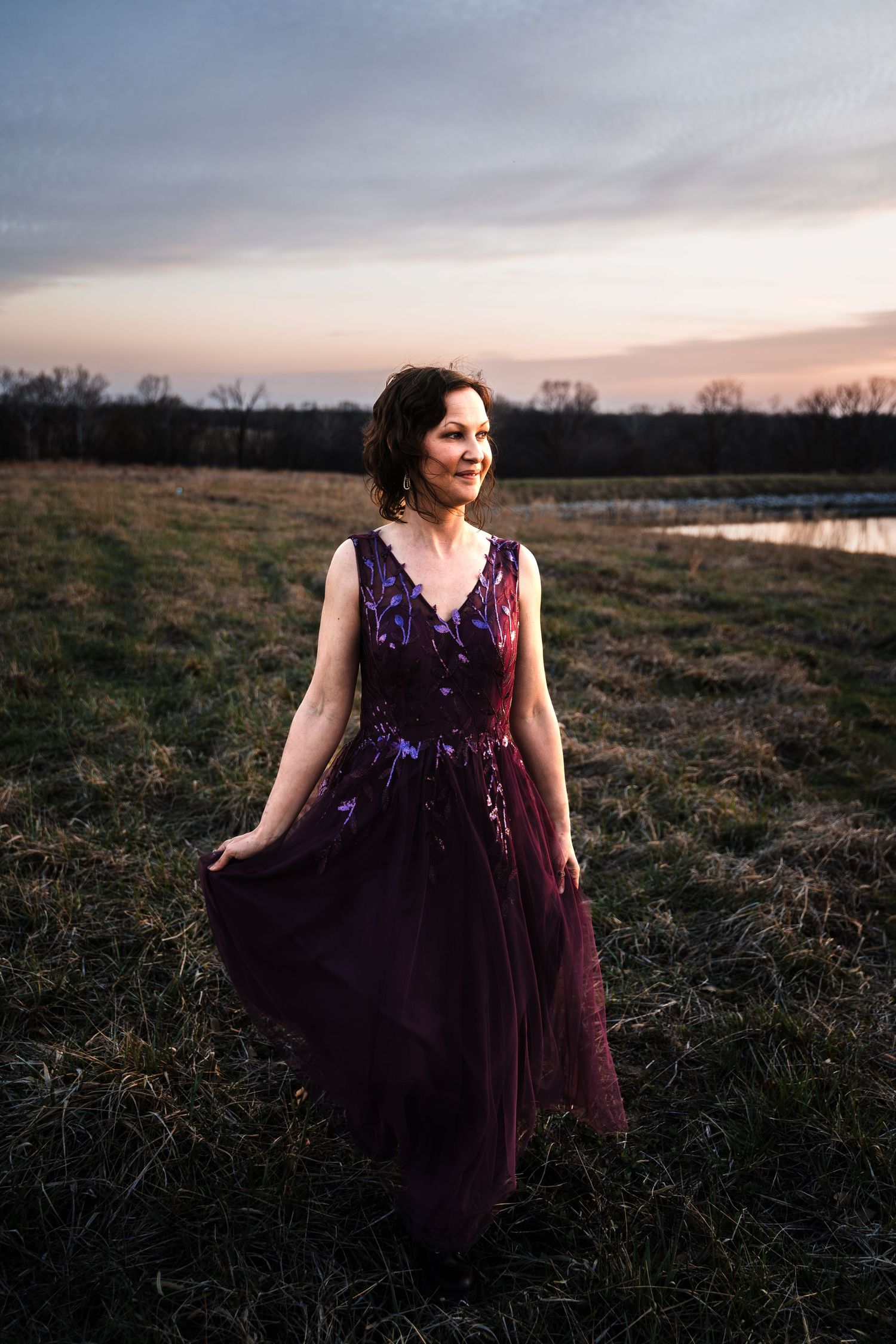 Woman in elegant purple tulle gown stands in grassy field during golden hour sunset.