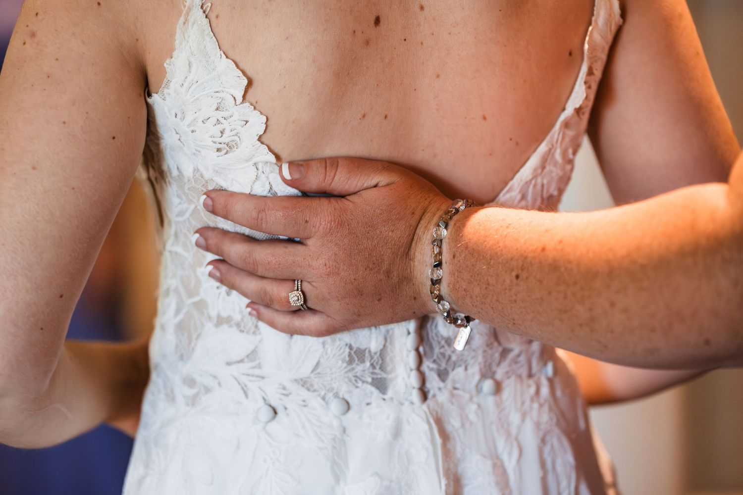 Close-up of a bride in lace wedding dress being helped by a woman wearing a bracelet and ring.