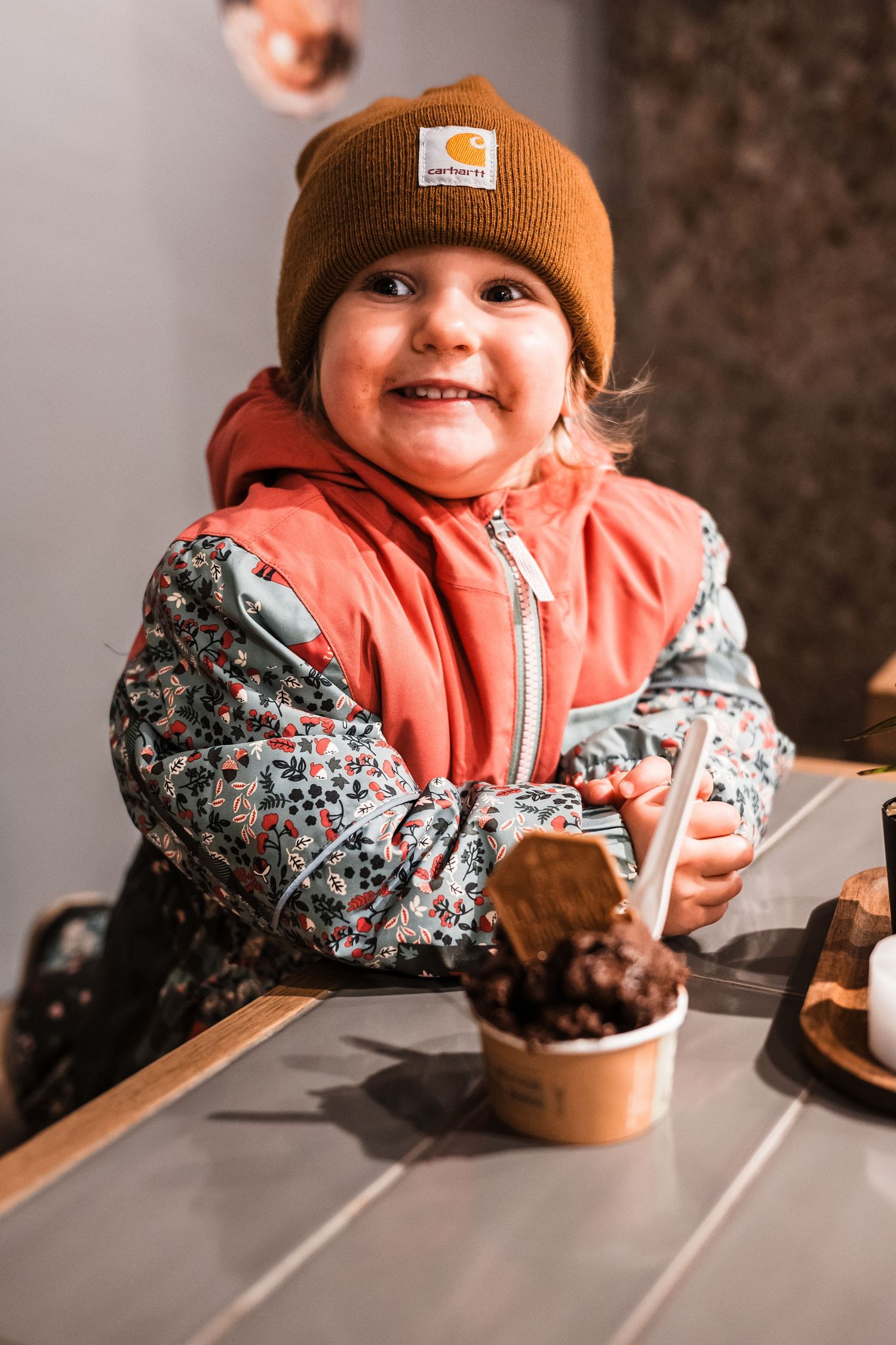 Small child in orange beanie and floral print jacket enjoys chocolate cupcake at wooden table in sequence of photos.