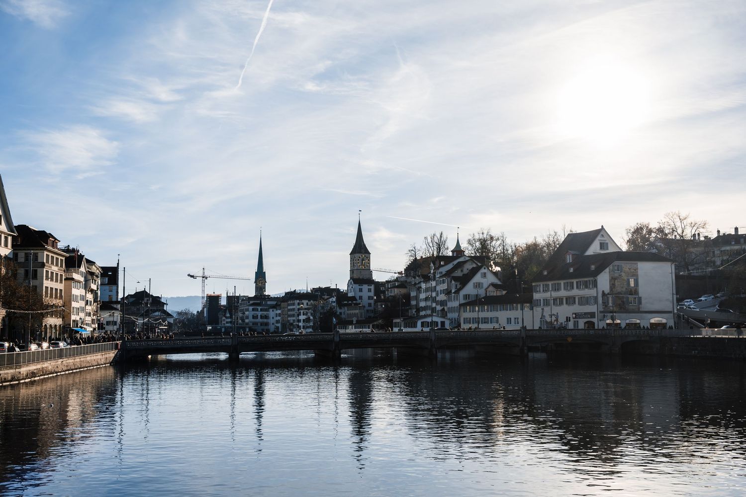 Panoramic cityscape view of a waterfront town with church spires and buildings reflecting in calm waters at dusk.