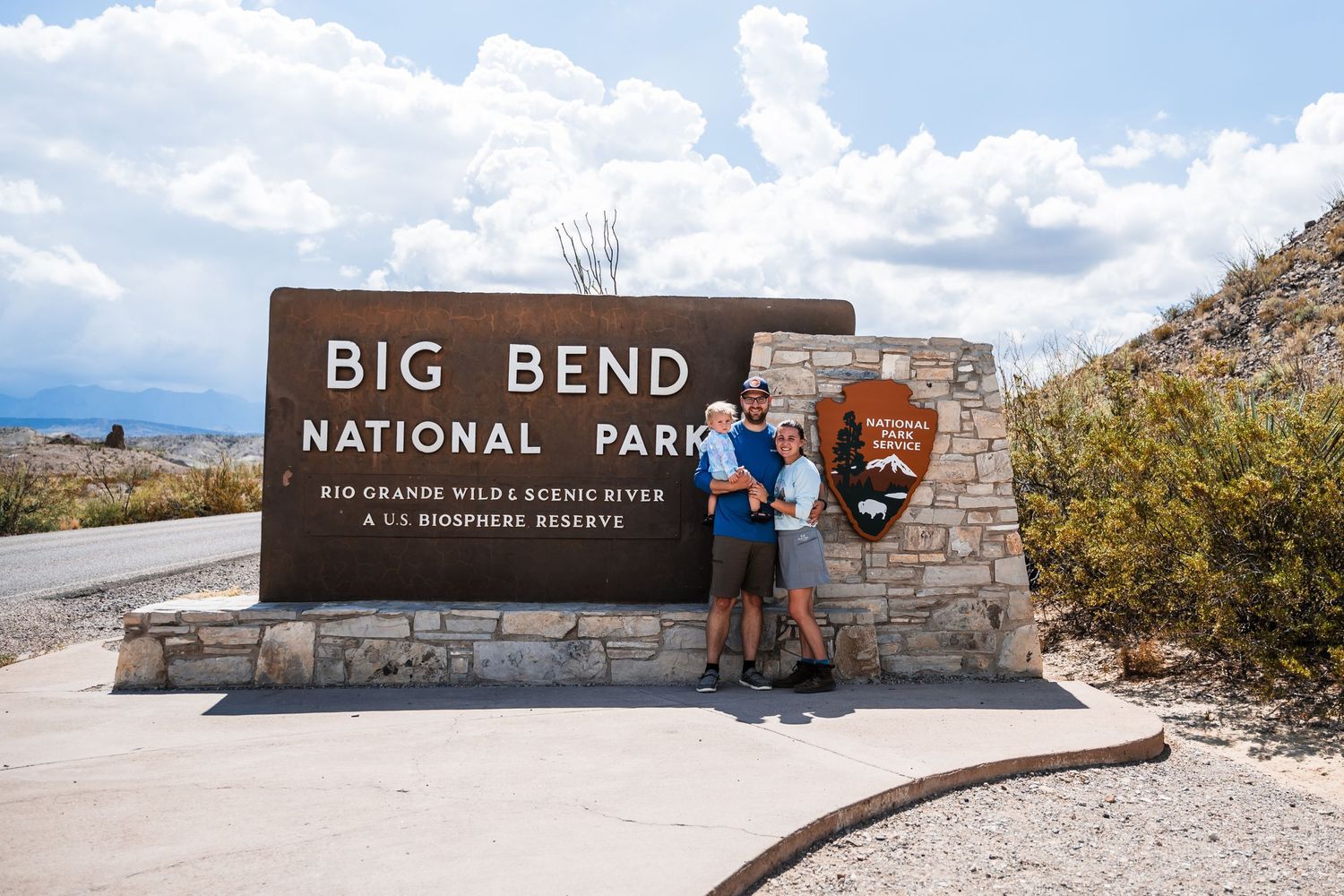 Visitors pose by the entrance sign at Big Bend National Park on a sunny day with mountains in the background.