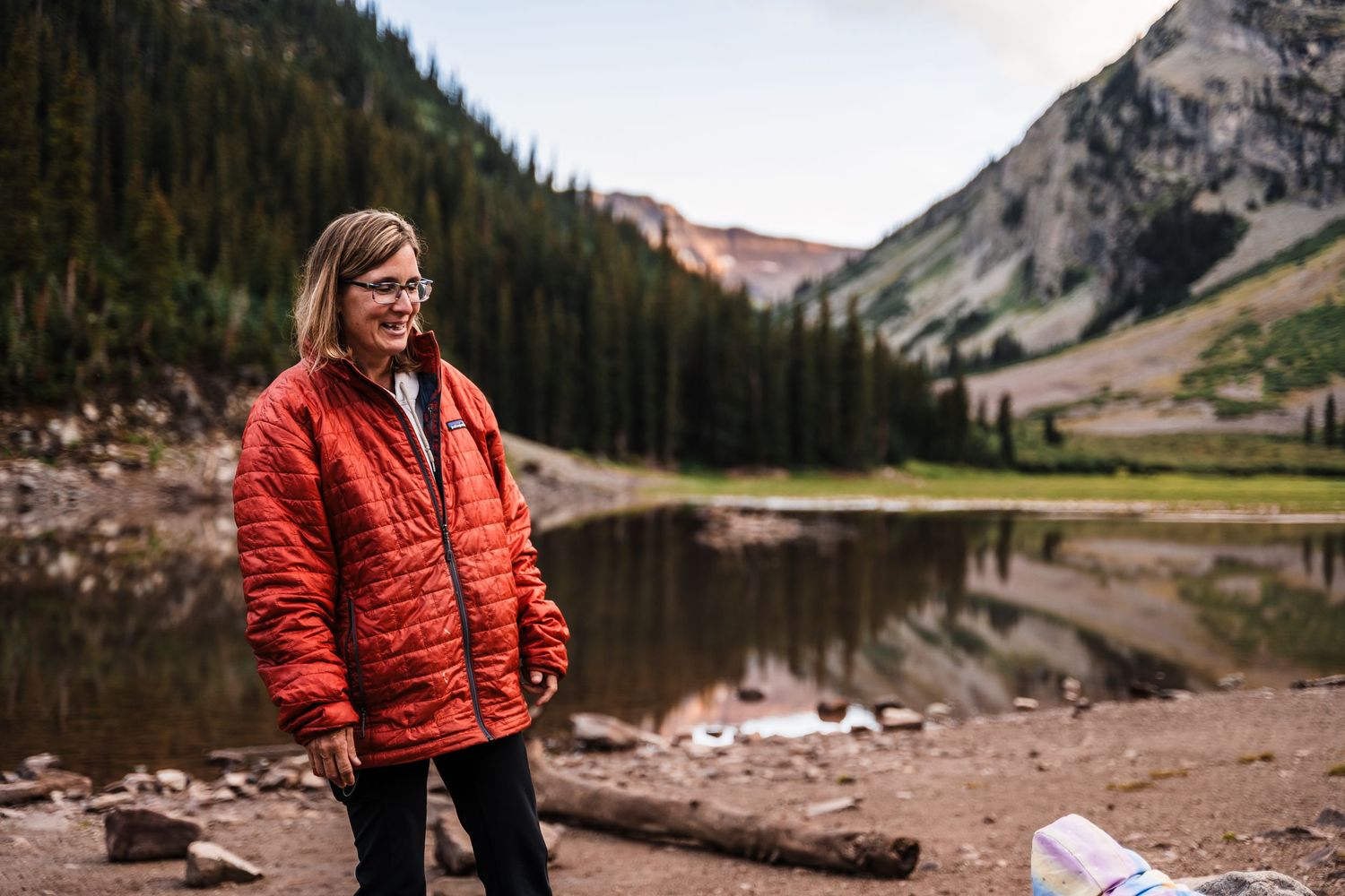 Hiker in red puffy jacket stands by mountain lake surrounded by evergreen forest and rocky peaks at dusk.