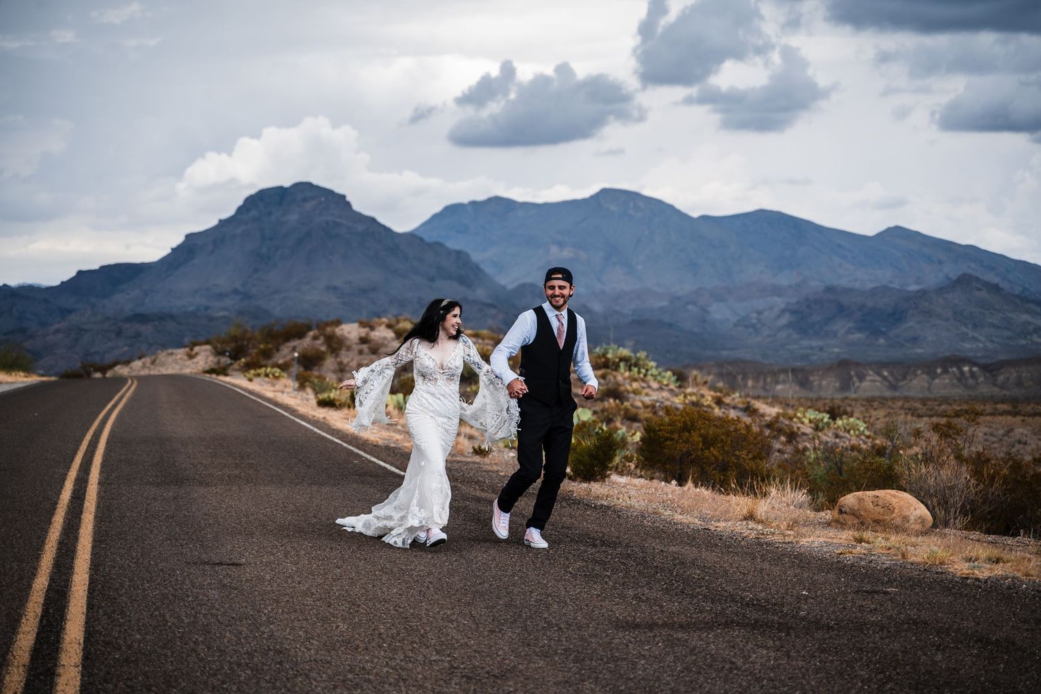 Couple walking on desert road with dramatic mountain landscape in background.