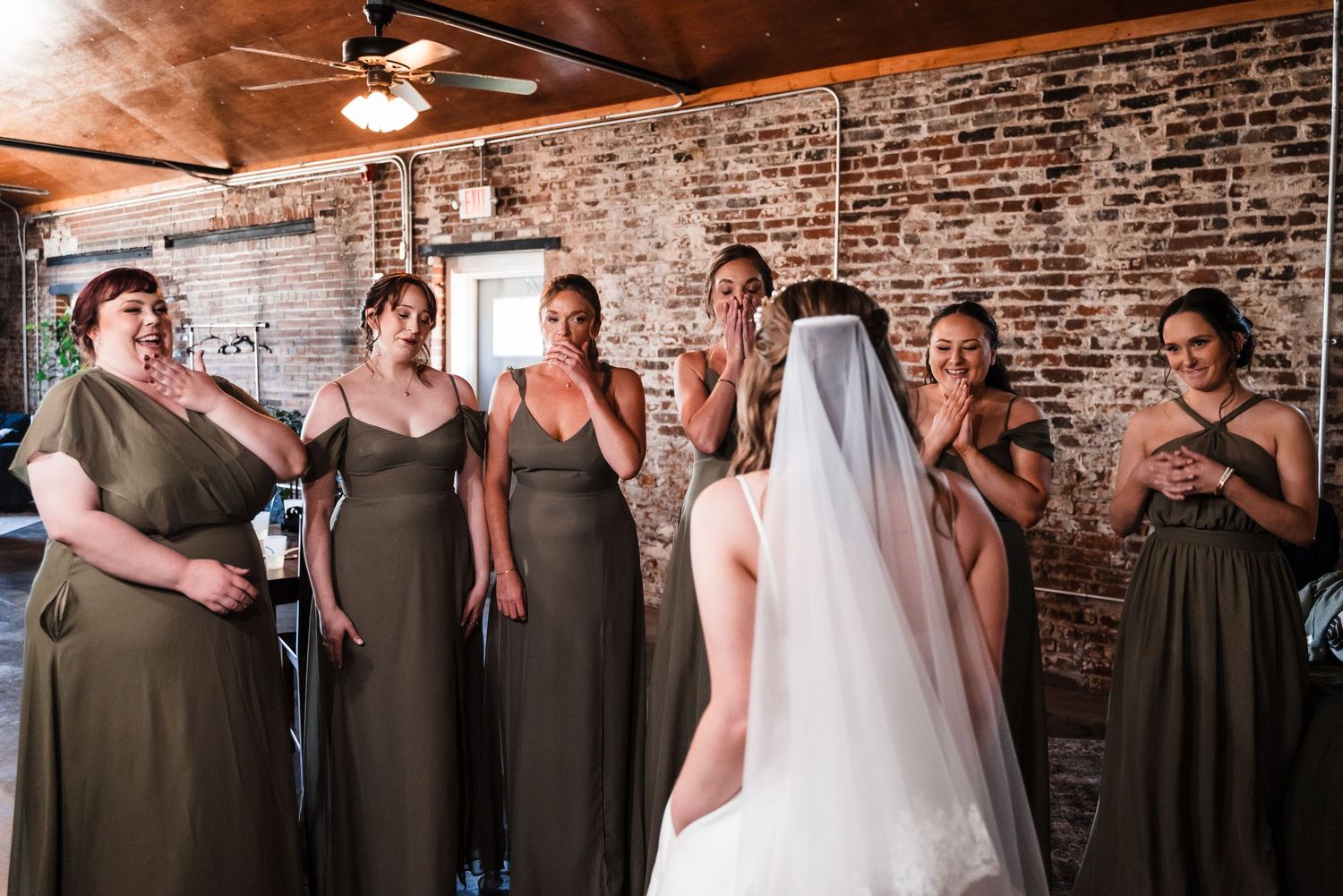 Wedding party in olive green dresses sharing an emotional moment in a rustic brick-walled venue.