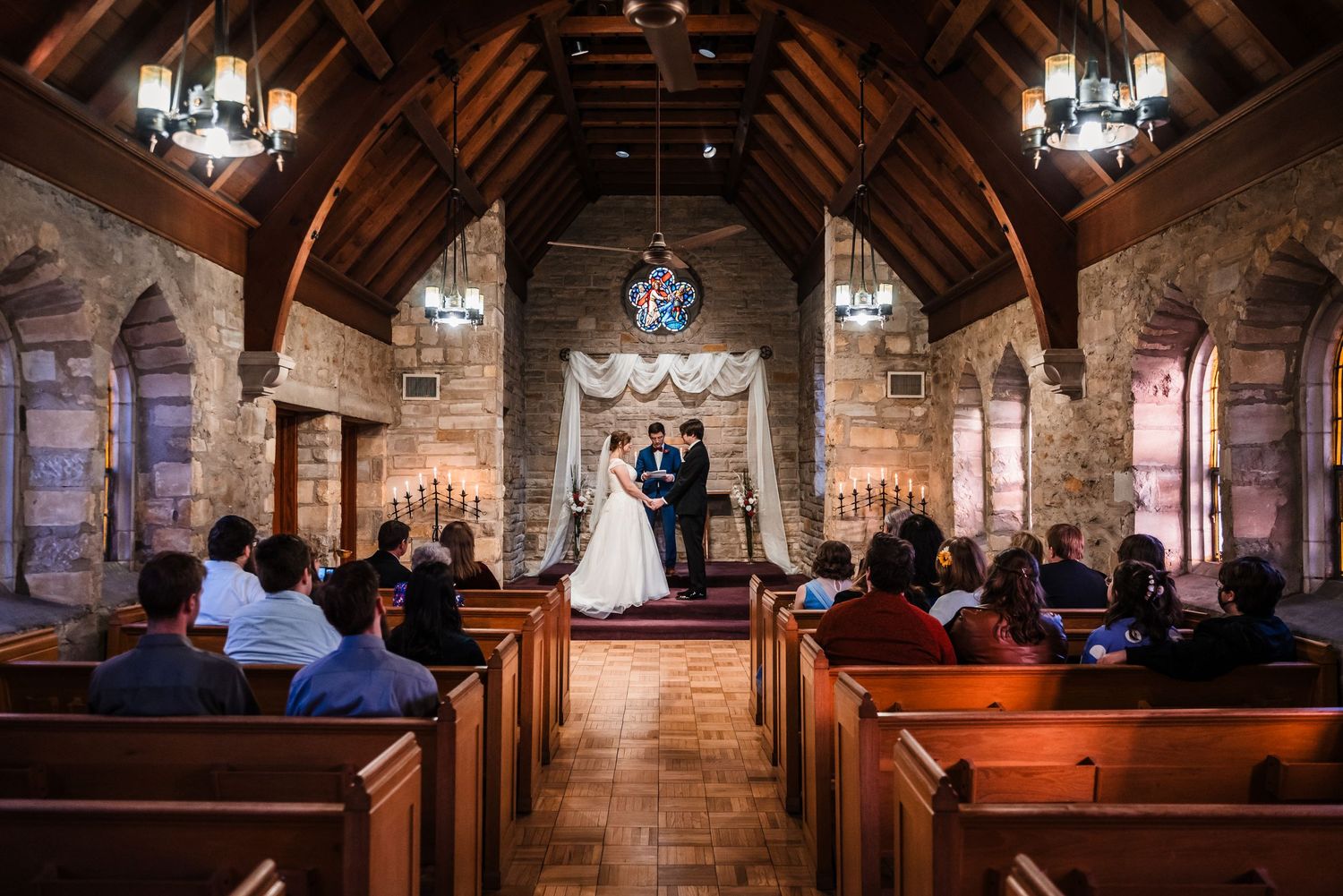 A stone chapel wedding ceremony with exposed wooden beams and stained glass windows illuminated by hanging chandeliers.