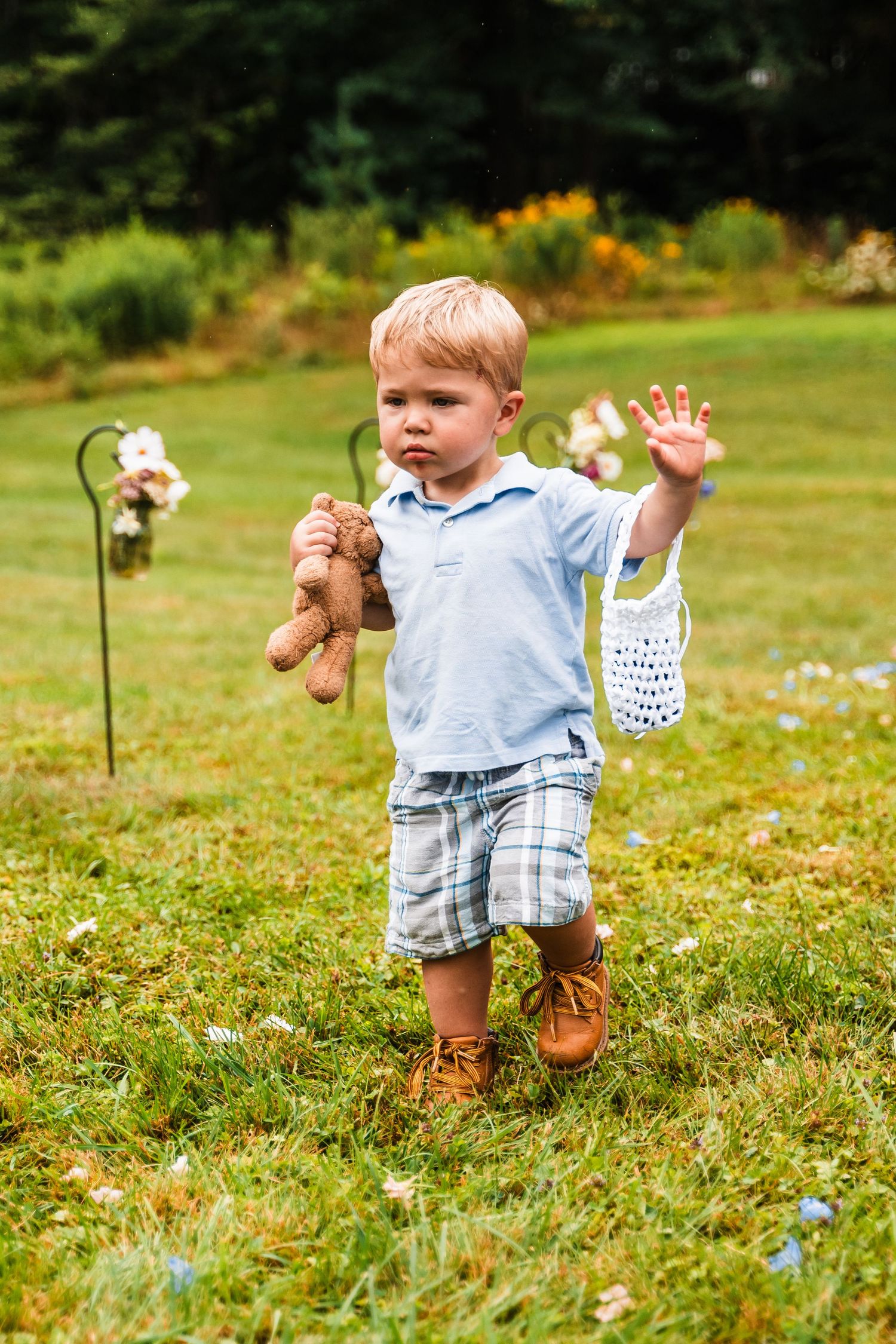 Toddler in blue shirt holds teddy bear while walking through garden setting.
