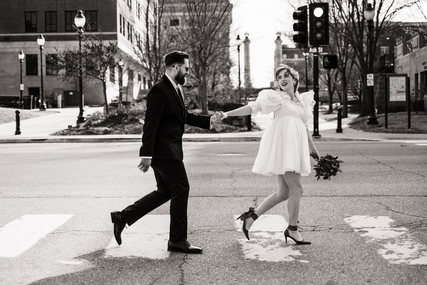 Black and white photo of a couple dancing joyfully across a crosswalk in formal attire.