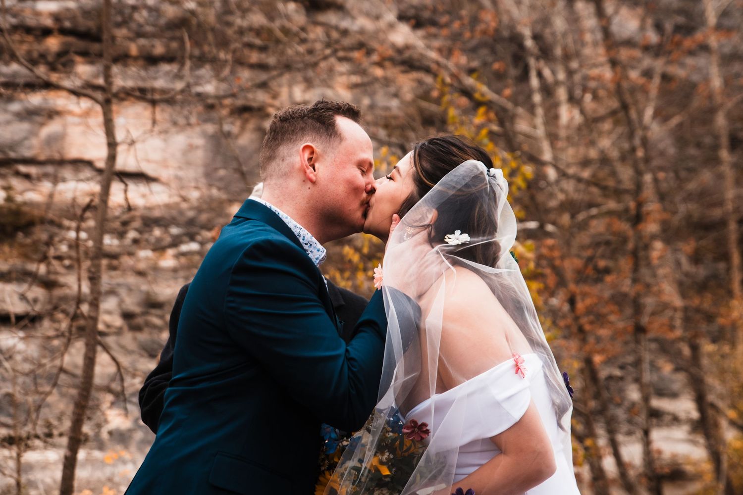 Romantic first kiss as newlyweds during fall outdoor wedding with rocky backdrop.
