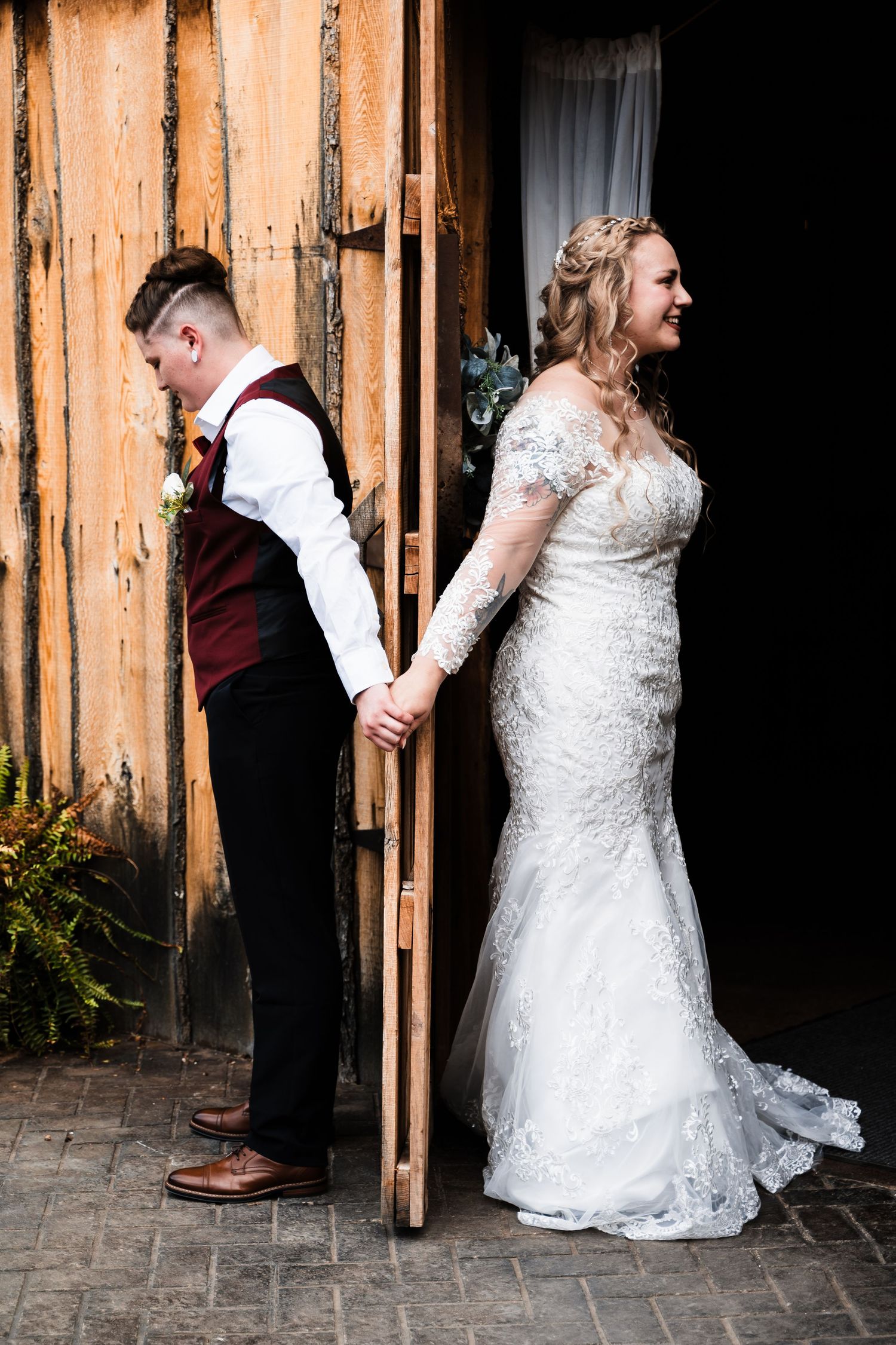 Artistic wedding photo with couple holding hands on opposite sides of rustic wooden wall.